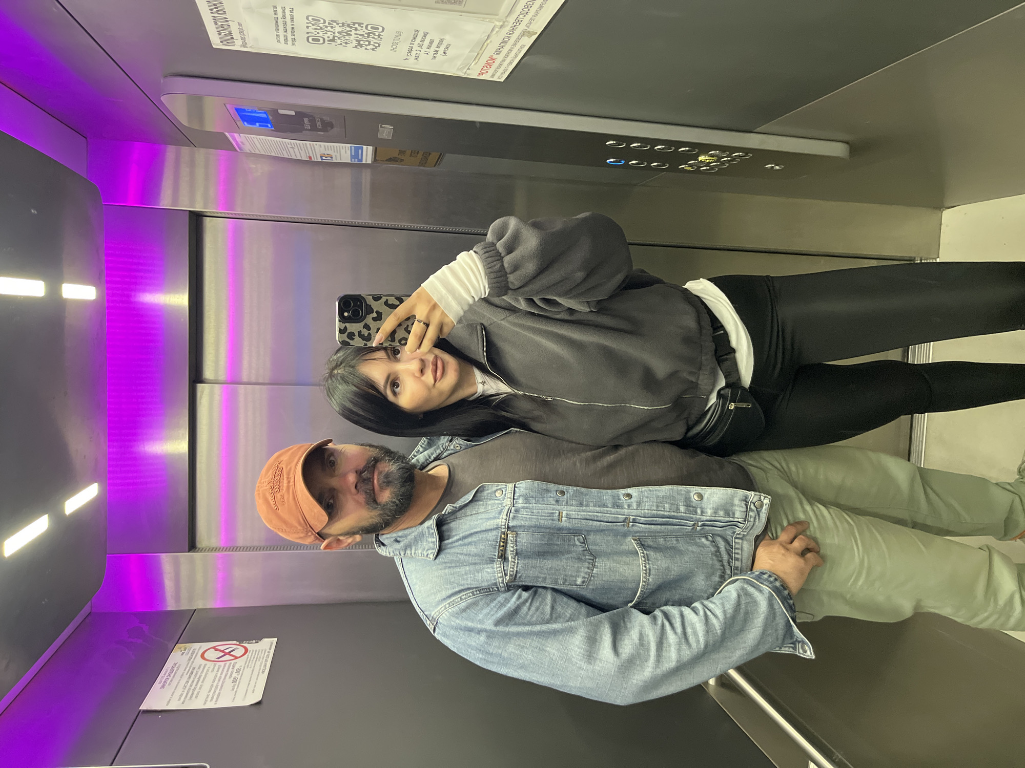 A man and woman are taking a selfie in an elevator. The man is wearing a denim jacket and an orange baseball cap, and the woman is wearing a black jacket and black pants. The elevator has purple lighting. A sign with Cyrillic text is visible on the elevator wall. Based on the sign and information available from image crops, it is possible that the location is Sochi, Russia.