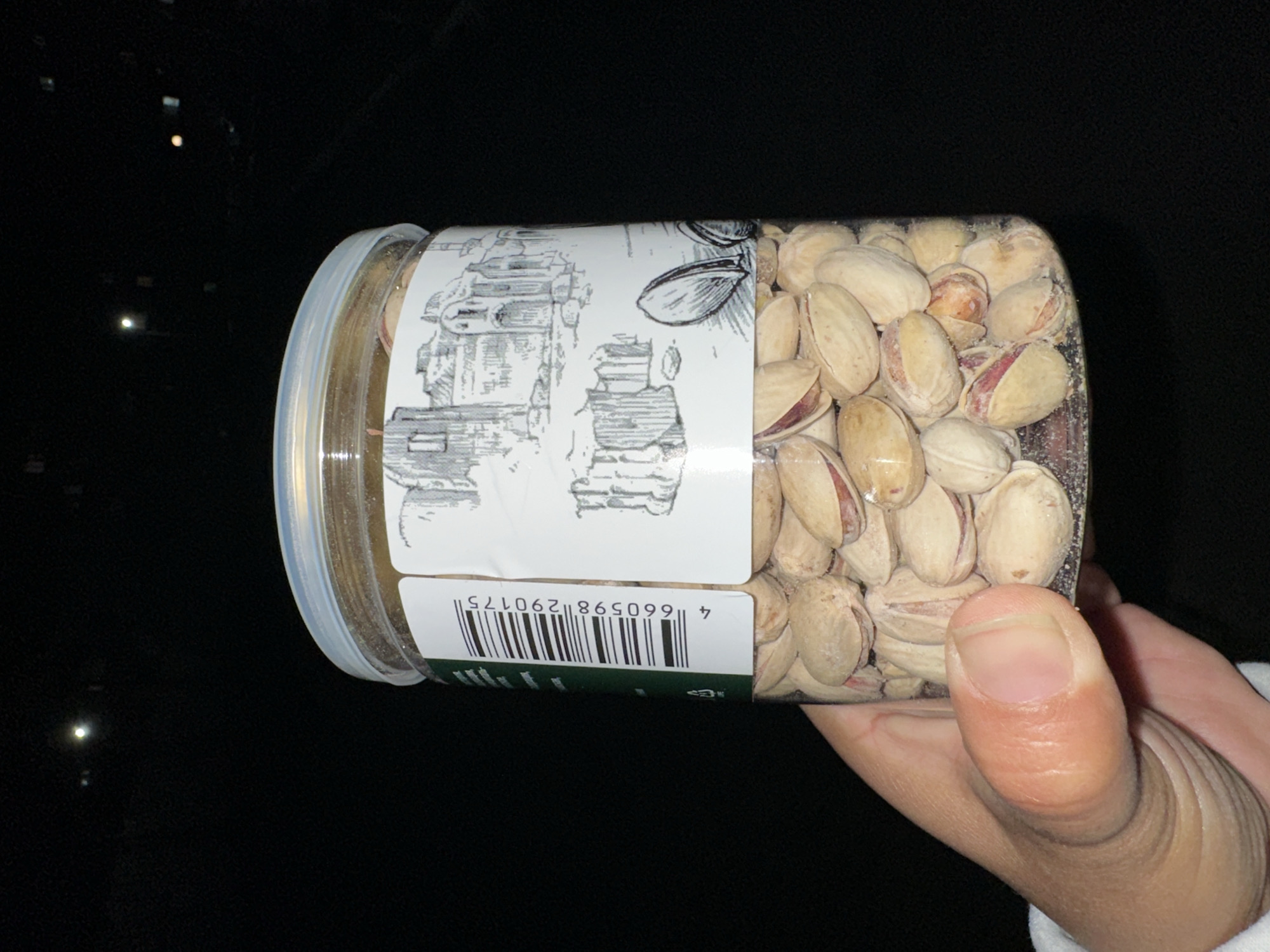 A hand holds a clear plastic container filled with pistachios against a dark background. The container has a white label with black line drawings and a barcode. The barcode number is 4 660598 290175. There are distant lights visible in the background.