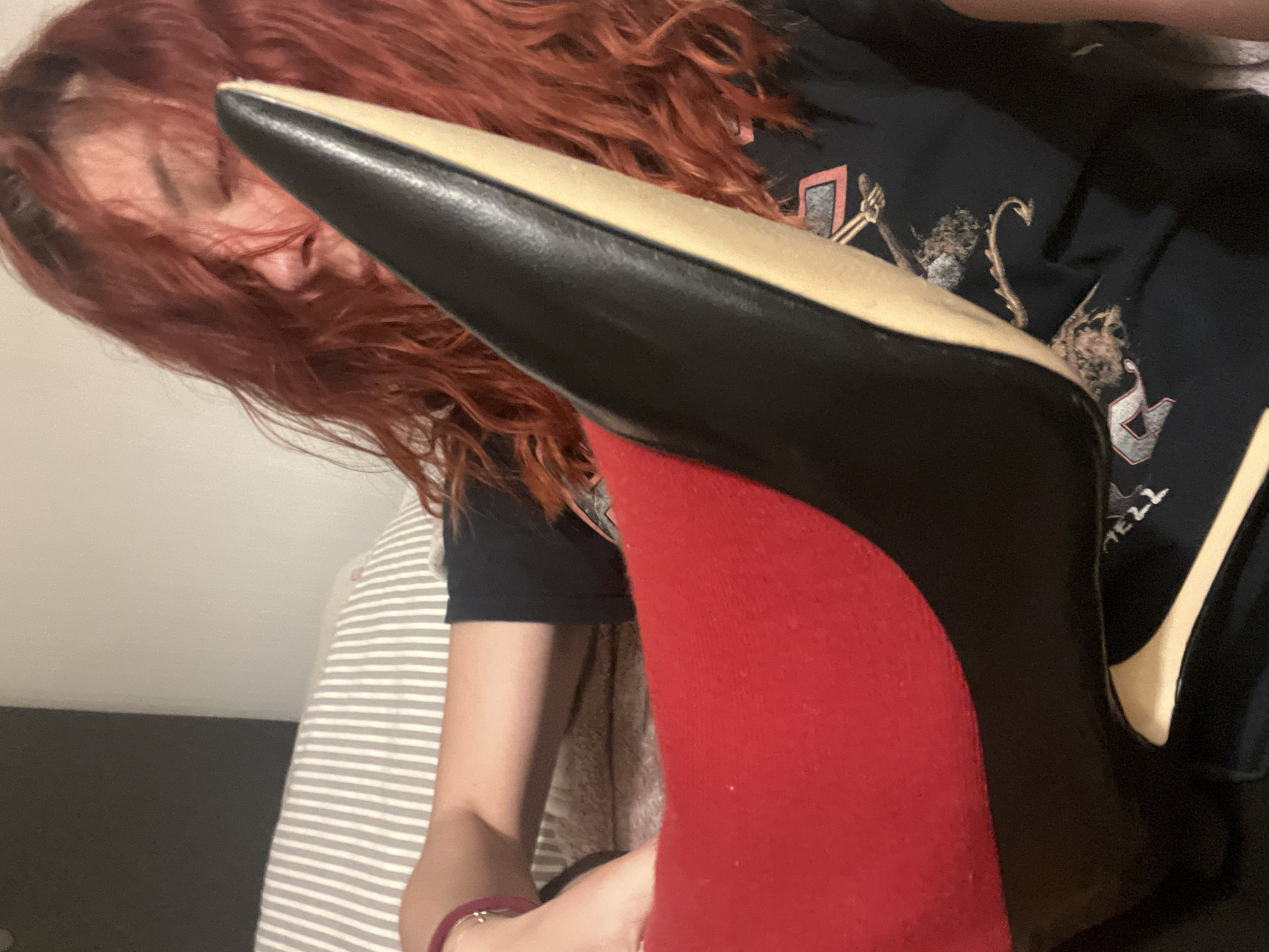 A person with red hair and wearing a black t-shirt is holding a black high-heeled shoe with a red sole. The background shows a striped surface and a dark grey area. The location is not discernible from the provided image.