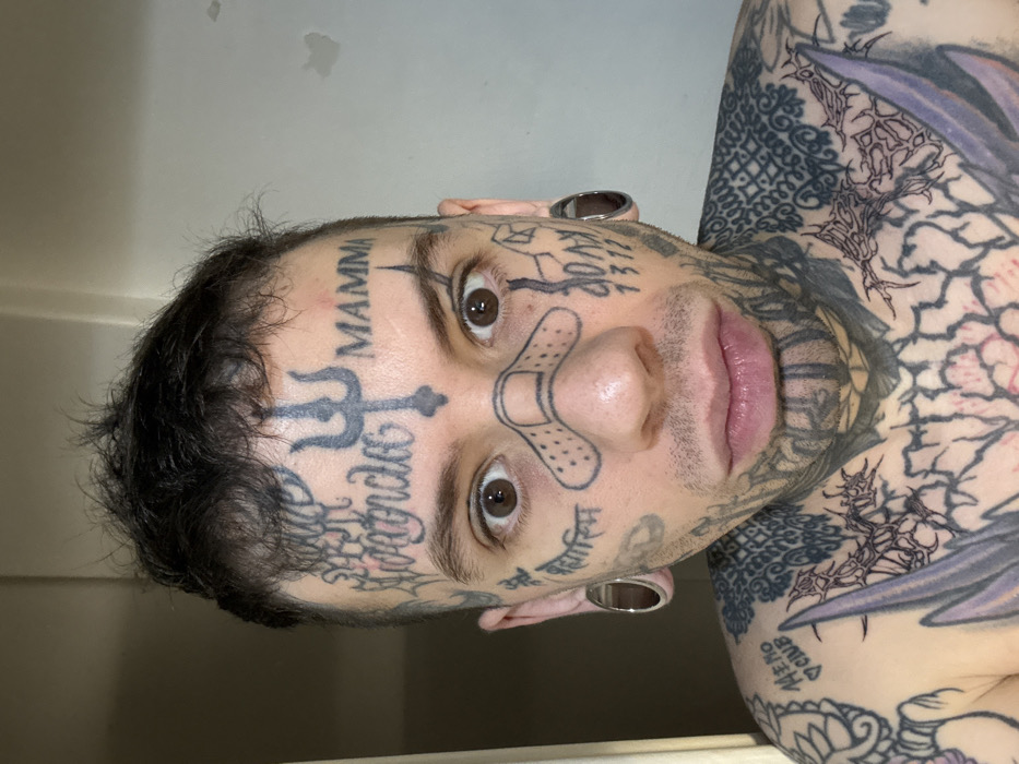 The image depicts a close-up, frontal portrait of a single individual, presumably male, with extensive facial and upper torso tattooing. The person is situated indoors, likely in a residential setting within Buenos Aires, Argentina, as indicated by a plain, light-colored wall with minor imperfections visible in the background.
The individual has short, dark, somewhat disheveled hair. Their face is heavily tattooed with black ink designs, including a trident symbol on the forehead, the word "MAMMA" to its right, and cursive script (partially visible, possibly "Alejandro") to its left. A bandage-shaped tattoo is positioned across the bridge of the nose. Various other symbols and text, such as "1312" and script resembling Devanagari, are present on the cheeks and jawline. Large, metallic-rimmed ear gauges are inserted into both earlobes. The individual has wide, brown eyes that are open and directed straight at the viewer, maintaining a neutral expression. The upper chest and shoulders are also extensively tattooed with intricate black ink patterns, including a large, stylized design across the sternum and a smaller tattoo reading "MEMO CINE" on the left collarbone area. The individual appears shirtless. No direct interactions with others are visible, suggesting a self-portrait or posed photograph.