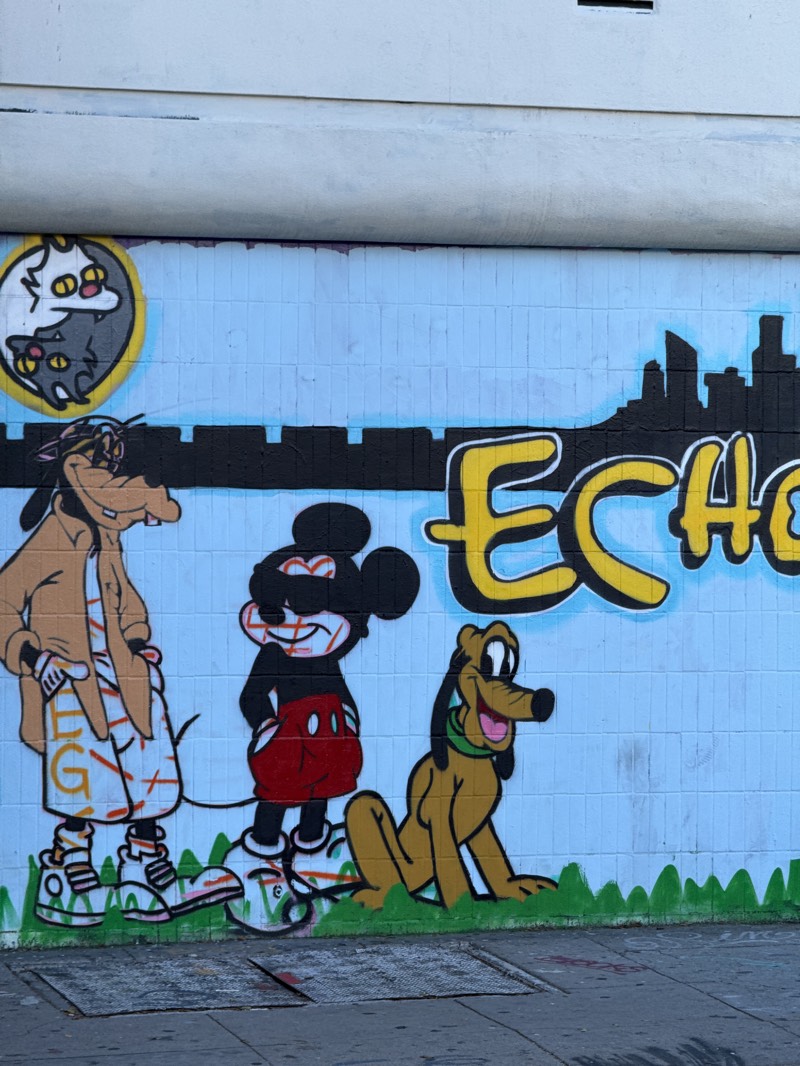 This is a close-up shot of a mural on a light blue brick wall. The mural features cartoon characters. On the far left is Goofy, wearing a brown jacket and white shorts with colorful graffiti. To Goofy's right is Mickey Mouse wearing a black shirt, red shorts, and white shoes. Mickey's face is covered in red graffiti. To the right of Mickey is Pluto, a yellow dog with a green collar, sitting on a patch of green grass. Above Goofy is a circle with two cats in the yin-yang position. Above Mickey is part of a black city skyline and graffiti letters. In the bottom of the image is the edge of a sidewalk. The mural is located in Los Angeles, United States.
