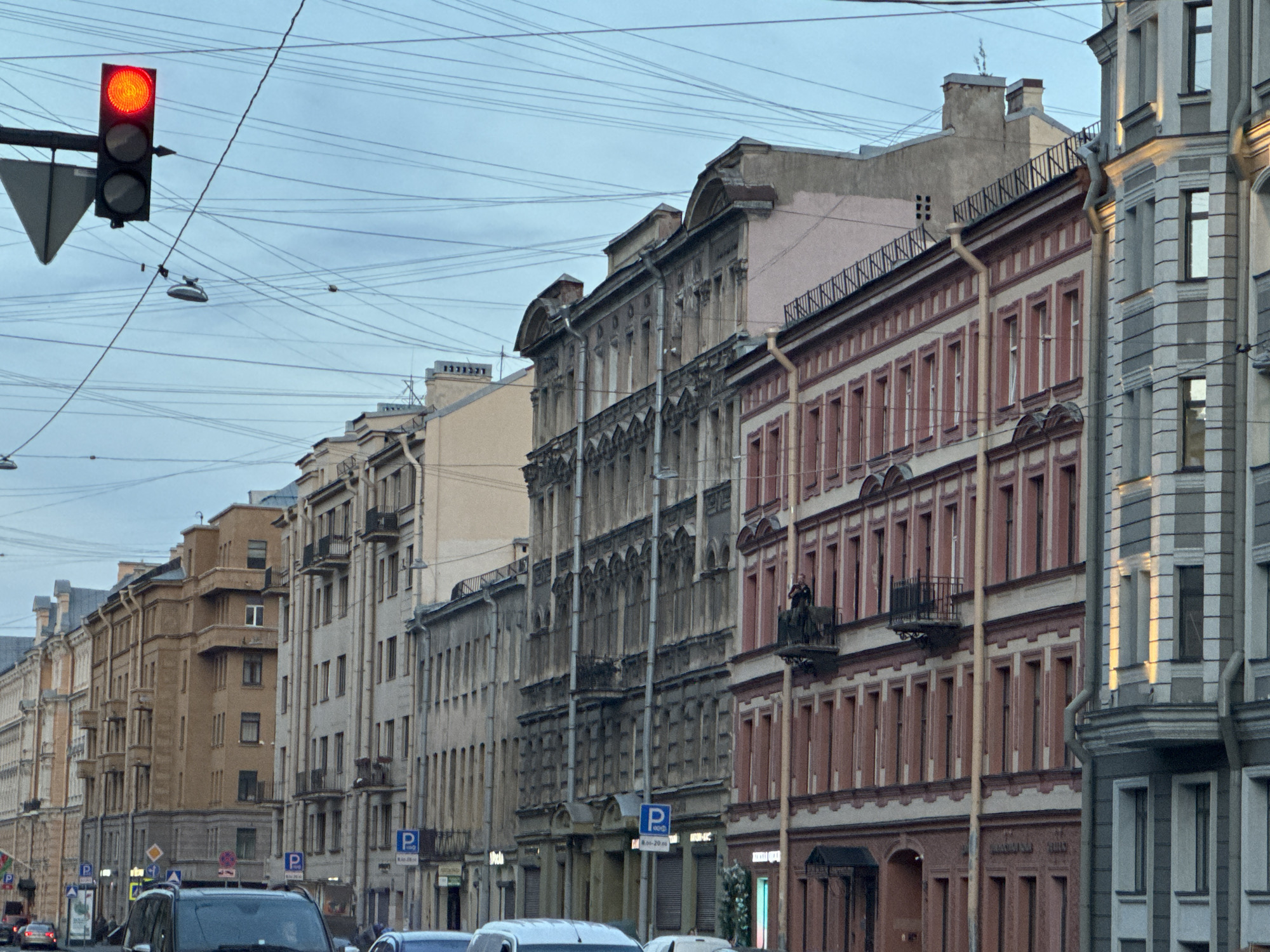 This image captures a street scene in the historic center of Saint Petersburg, Russia. The perspective is from street level, looking down a thoroughfare lined with multi-story classical and eclectic buildings under an overcast, light blue-grey sky.
In the upper left, a black traffic light fixture displays a red signal. A dense network of black overhead power lines, likely for trolleybuses or trams, crisscrosses the sky above the street. Suspended streetlights are also visible among these wires.
On the far right, a prominent multi-story building features a distinctive facade with alternating vertical sections of lighter and darker grey stone or stucco. Adjacent to it on the left is a multi-story building with a reddish-brown lower facade and lighter, cream-colored upper sections. This building has numerous uniformly spaced rectangular windows. On an upper floor, a black metal balcony extends from the building, where a single person is visible standing and looking towards the left, appearing to hold a device, possibly a camera or phone.
Further down the street, a darker, ornate multi-story building with a greyish facade displays multiple windows topped with decorative arch-like elements. Vertical drainpipes run down its exterior. A series of additional multi-story buildings, varying in architectural style and color (including beige, brown, and lighter grey), extend into the distance, some featuring balconies.
At street level, parts of several vehicles are visible, including the dark hood of an SUV in the immediate foreground. Blue "P" parking signs, indicating parking regulations, are positioned along the sidewalk; one clearly displays "8.00-20.00" and Cyrillic text underneath. The overall scene depicts a busy urban environment during daytime with soft, diffused light.