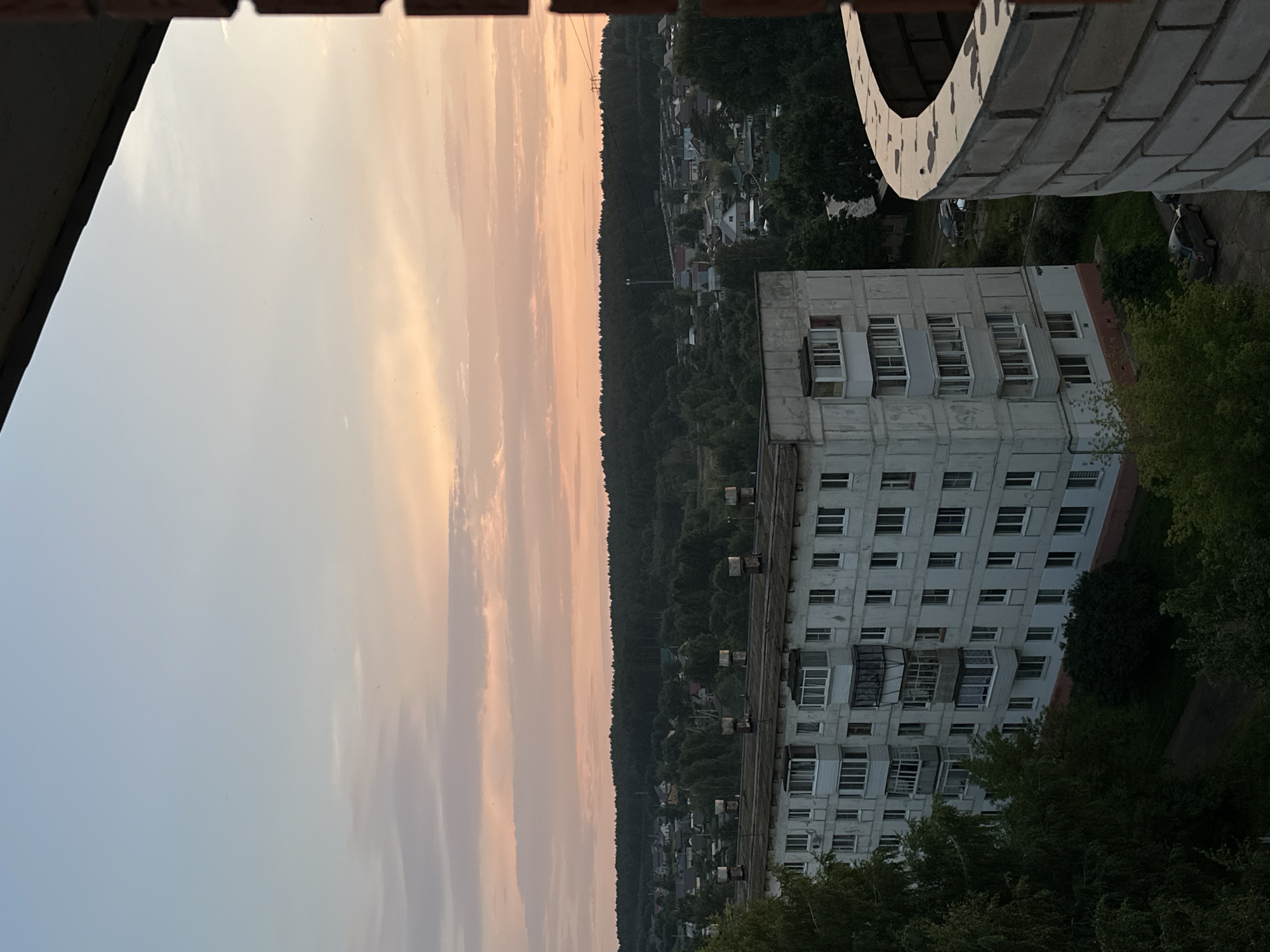 The image depicts a residential building in the city of Chaikovsky, Russia, at dusk. The building is a multi-story, concrete structure with numerous windows and balconies. The sky above is filled with soft, pastel clouds, transitioning from a pale blue at the top to hues of pink and orange near the horizon. A dense line of trees forms a dark silhouette against the colorful sky, behind which a scattering of smaller houses and buildings is visible, suggesting a suburban or semi-rural area. In the foreground, a curved brick structure, likely a balcony or parapet, partially obscures the view. The overall impression is one of a quiet evening in a residential district.