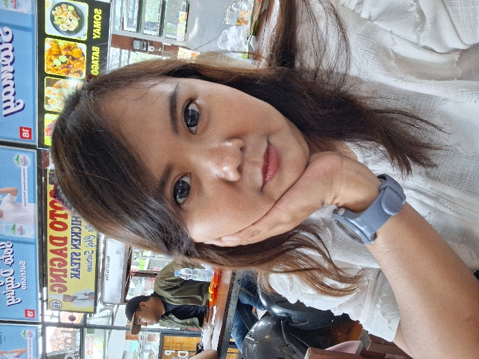 A woman with long brown hair and a white shirt is shown in a close-up shot. She rests her face on her hand, wearing a gray watch. The background reveals a food stand with a menu board advertising "SOTO DAGING" or meat stew. There is another person wearing a cap and jacket seated in the background, possibly at a table in the eatery.