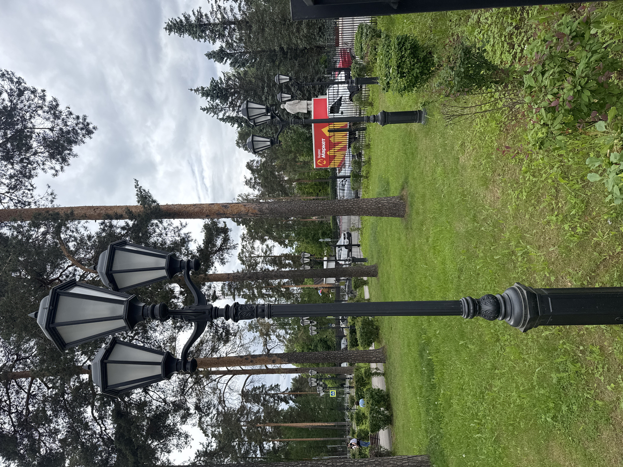 In a city park setting on a cloudy day, a black, ornate lamppost with three lights stands in the foreground on the left. Behind it, a path winds through a grassy area with manicured bushes and tall pine trees. Further in the background, a black metal fence encloses a street with parked cars and a red truck featuring the "Yandex Market" logo. A statue of a person stands on top of the truck. Several other lampposts are visible along the path. On the left side of the image, two individuals are seated on a bench, with one person standing. Another person is walking on the path. The overall environment suggests a public outdoor space with urban elements.
