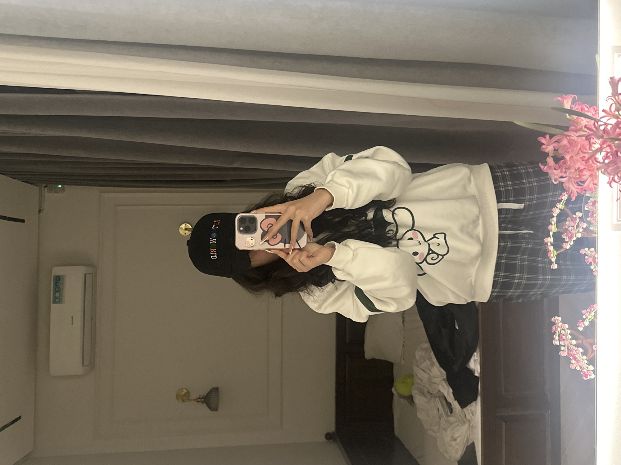 A person is reflected in a mirror. The person is wearing a black hat, a white sweatshirt with a cartoon image, and plaid pants. They are holding a phone with a patterned case, presumably taking a selfie. The reflection shows a bedroom interior. The background includes a window with a curtain, a wall-mounted air conditioner, and a bed. The location is not discernible from the image.