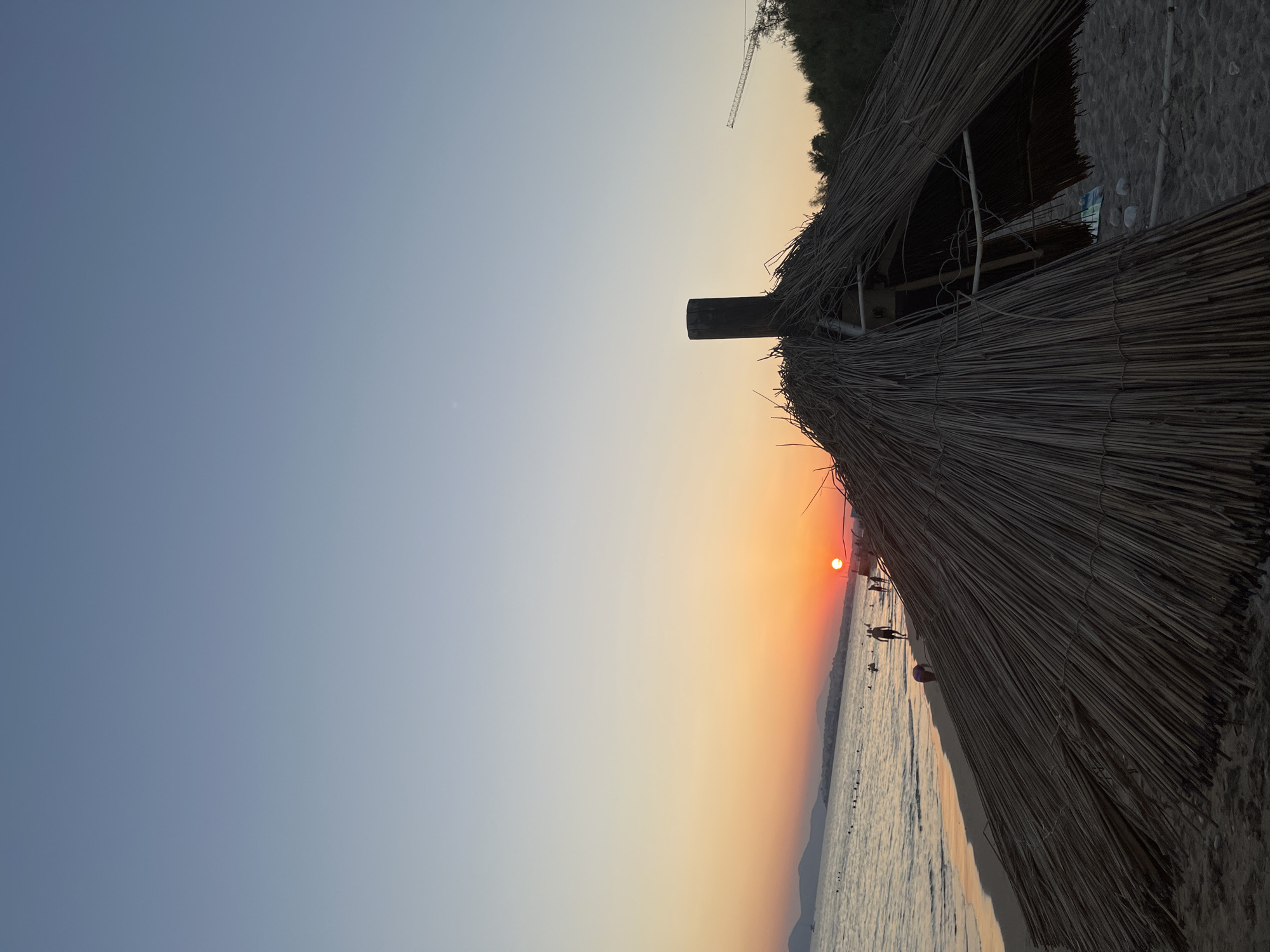 This is a photograph of a beach at sunset in Paleo Faliro, Greece. The sun is setting over the sea, casting an orange glow across the sky and water. In the foreground, there is a thatched umbrella, partially collapsed. People are visible on the beach and in the water, some swimming and others walking along the shore. The water is calm and the sky is clear. In the distance, mountains can be seen along the coastline. A construction crane is visible on the right side of the image, against the horizon. The scene captures a peaceful evening at the beach.
