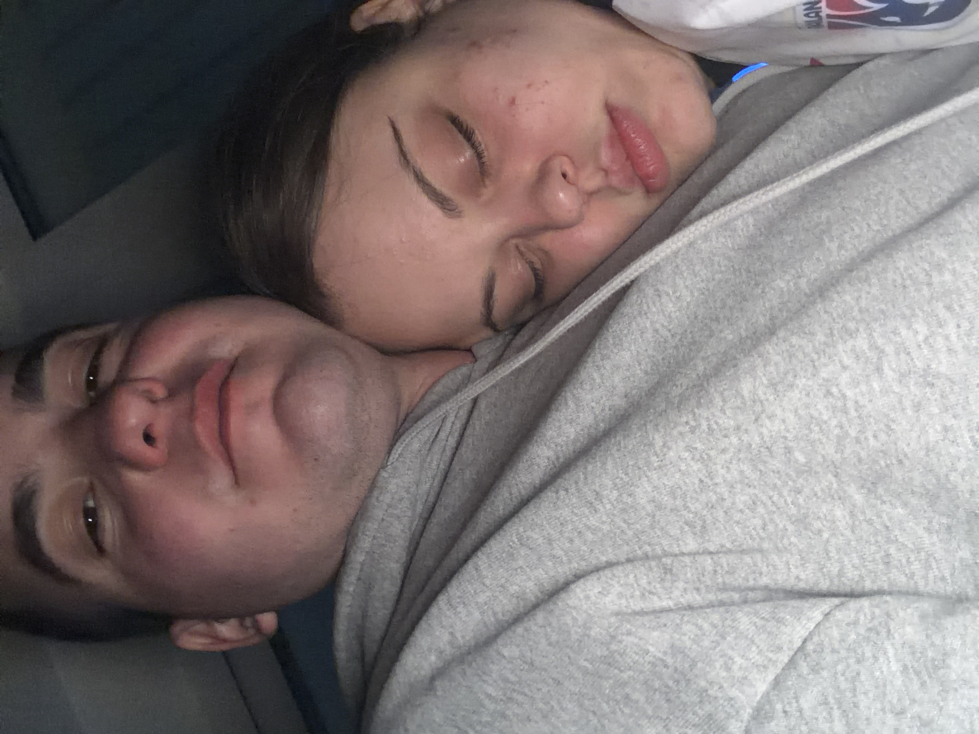 An intimate, close-up shot captures two individuals, a man and a woman, cuddling or resting together in what appears to be an indoor setting in Varna, Bulgaria. The image is taken from a slightly upward, diagonal angle, emphasizing their faces and shared space.
The woman, with dark hair, is nestled asleep against the man's chest. Her eyes are closed, her face is relaxed, and she has visible acne marks on her forehead. Her lips are slightly parted.
The man is positioned beneath her, looking towards the camera with a soft, relaxed expression. His eyes are open, and his face shows a faint blush. Both individuals are wearing light grey hooded sweatshirts, suggesting comfort and casual attire. The drawstring of the man's hoodie is visible on his chest.
Behind the woman's head, a white fabric item with blue and red markings is partially visible, showing the letters "MAN," likely part of a logo or text on a piece of clothing or blanket. The background is dark and out of focus, providing no specific details about the room but contributing to a cozy, dimly lit atmosphere. The overall mood conveyed is one of affection, comfort, and peaceful intimacy.