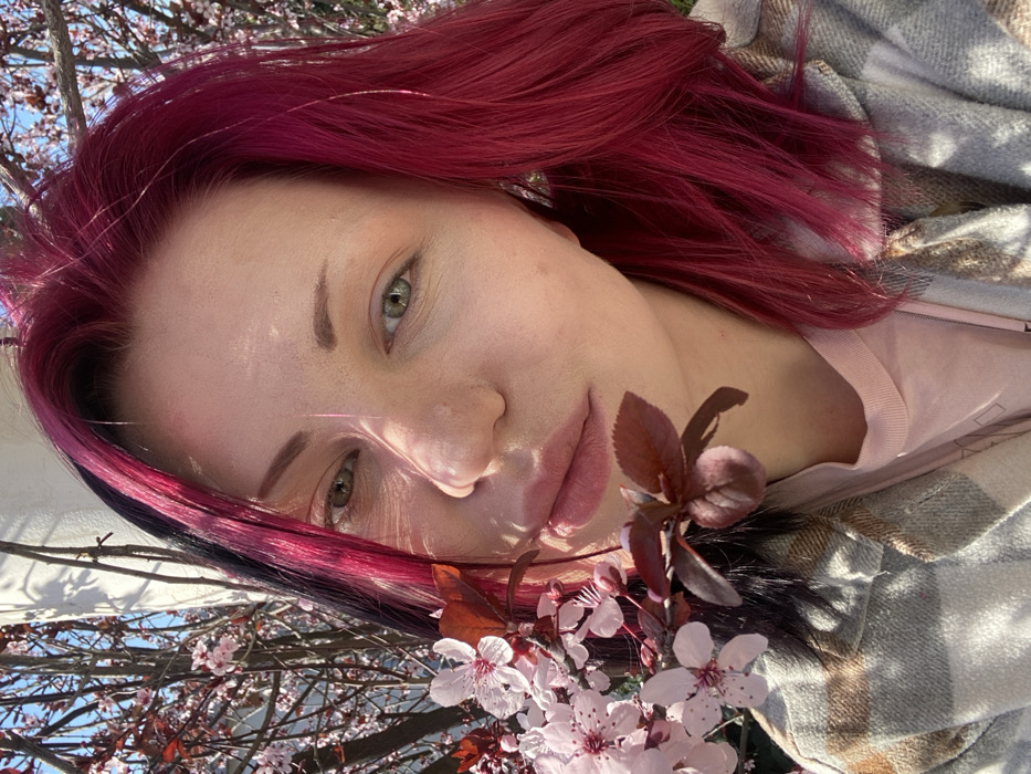 The image displays a close-up, outdoor portrait of an individual in Kunje, Montenegro. The subject is an adult female with fair skin, green eyes, and prominent pink-magenta hair, featuring a darker, possibly black or dark brown, streak on the left side. Her eyebrows are light brown and arched. Her lips are full and light pink. She wears a light-colored, possibly plaid or checkered, collared jacket over a light pink t-shirt. A metallic earring is visible on her right ear. A branch featuring light pink blossoms and dark red leaves is positioned diagonally across the lower left foreground, extending towards the subject's face and shoulder. Bright sunlight illuminates the scene, casting noticeable shadows on the subject's face and upper body. The background is blurred, showing a tree with similar light pink blossoms against a clear blue sky, with a light-colored architectural element partially visible in the upper left. The individual is looking directly towards the camera.