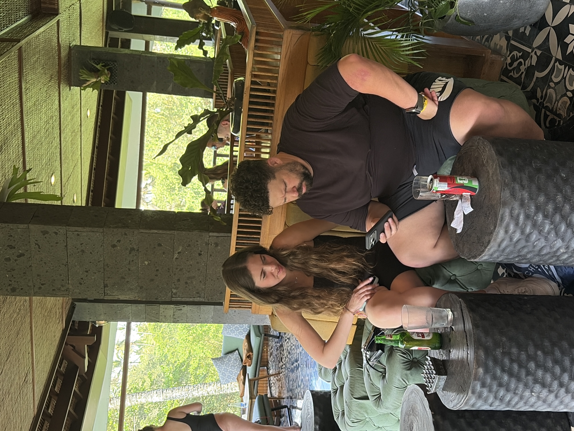 In Tegallalang, Indonesia, a woman with long brown hair and a black dress sits on a green tufted couch, looking at her phone. Next to her, a man with curly dark hair and a black t-shirt and shorts is also looking at his phone. They are seated in a lounge area with dark, textured cylindrical tables. On one table, there is a green bottle of beer and a glass of water, along with an ashtray. On another table, a can of Coca-Cola and a piece of white paper are visible. In the background, a woman in a black swimsuit stands near a sofa and a table with cushioned seating. The environment appears to be an open-air establishment with natural light filtering through lush green foliage and trees. The architecture features dark stone columns and a thatched ceiling.