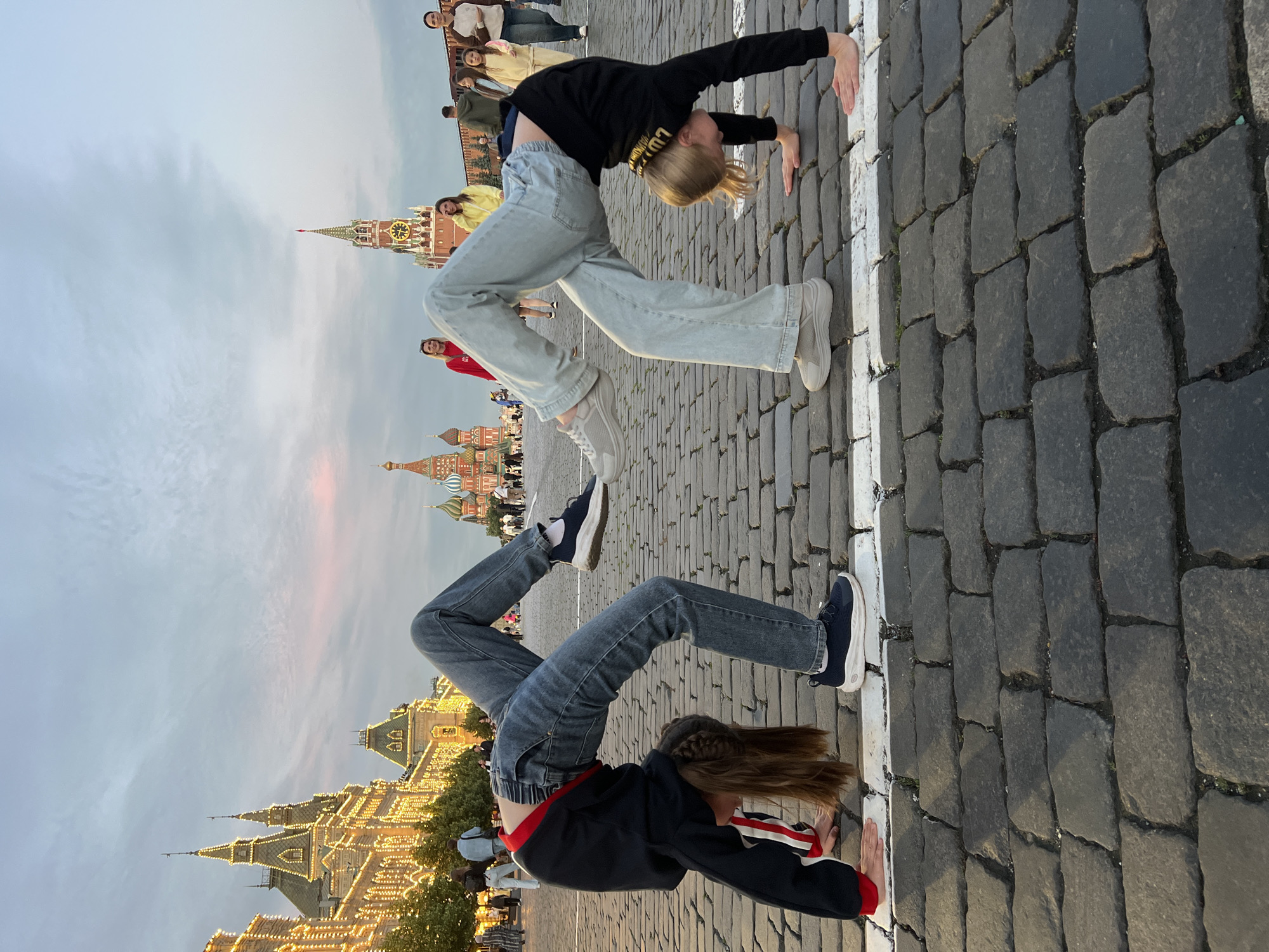 Two young individuals are performing a bridge pose on a cobblestone plaza in Moscow, Russia, with iconic architectural landmarks in the background. The person on the left, facing away from the camera, has dark hair braided and wears a black and red jacket with blue jeans and dark sneakers. The person on the right, facing the camera, has blonde hair and wears a black top with light blue jeans and light grey sneakers. Both are in a symmetrical, inverted bridge position, with their hands on the ground and their legs arched upwards.
In the background, to the left, is the GUM department store, illuminated by numerous lights, and beyond it, the towers of the Kremlin. To the right, the Spasskaya Tower, a prominent clock tower of the Kremlin, is visible. The sky above is overcast with hints of pink and orange from the setting sun. Other people are visible walking in the background, indicating a public space. The ground is paved with large, irregularly shaped cobblestones, with a white line demarcated on the ground between the two individuals. The overall scene captures a moment of athletic activity set against a historically significant backdrop in Moscow.