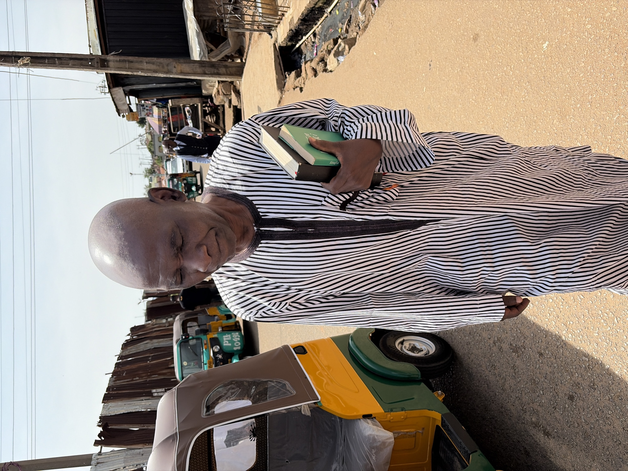A man with a shaved head, wearing a black and white striped robe, walks with two books in his right hand. He is on a paved street in Jos, Nigeria, with a yellow and green auto-rickshaw to his left. In the background, corrugated metal fences, market stalls, and other auto-rickshaws are visible under a clear sky. The man's gaze is directed downwards. The environment suggests a busy urban setting.