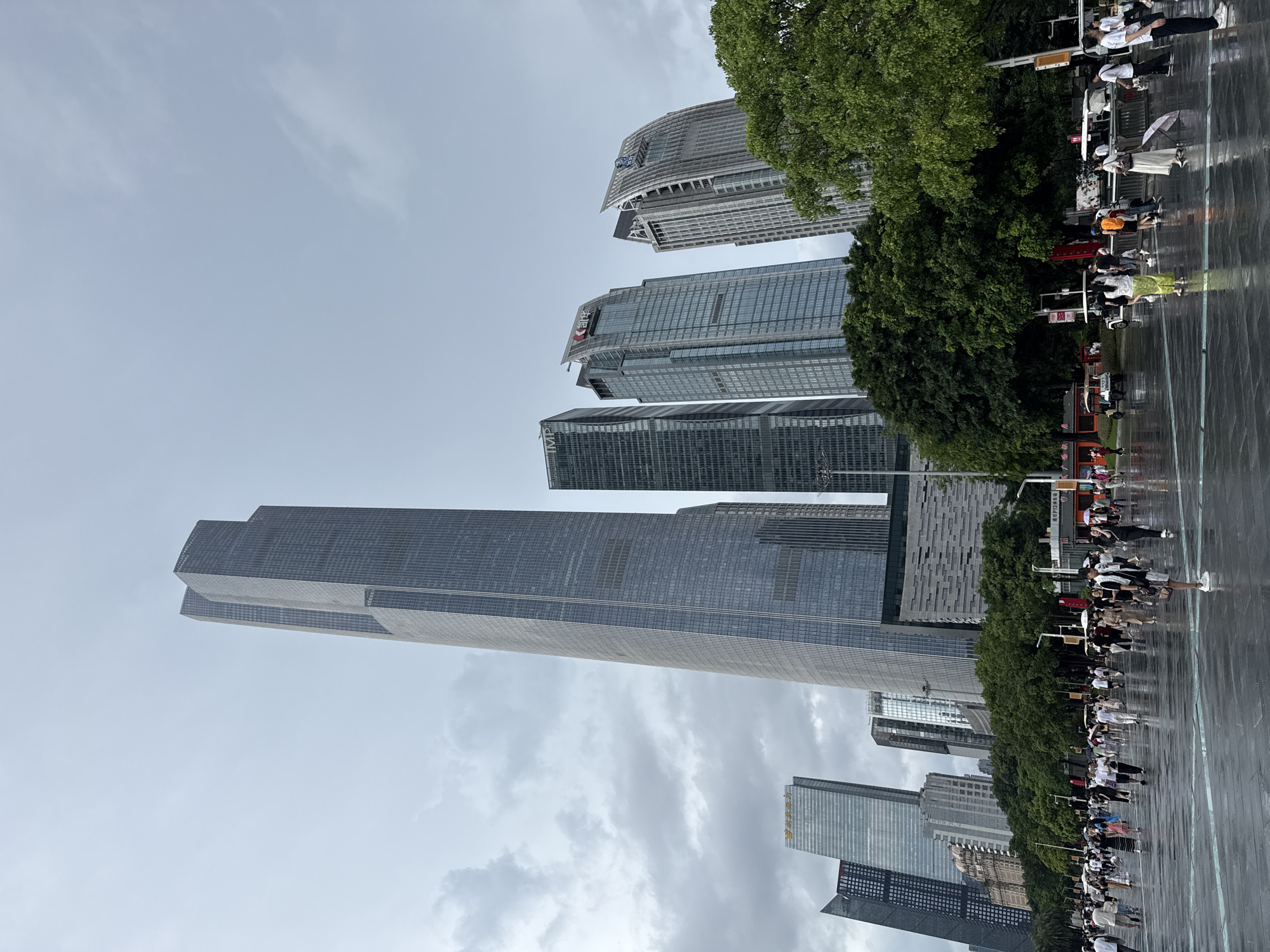 This image depicts a wide-angle view of a modern urban plaza in Tianhe District, China, under a cloudy sky. In the foreground, a group of people are walking on a wet, paved surface, suggesting recent rainfall. Several individuals are holding umbrellas. The plaza is bordered by a line of lush green trees. Beyond the trees, a cluster of imposing skyscrapers dominates the background, showcasing contemporary architecture with glass and metal facades. The tallest building, a slender, multi-faceted skyscraper, stands prominently in the center. Other buildings exhibit varying heights and designs, contributing to a dense urban skyline. The overall scene captures a bustling city environment, possibly after a rain shower, with a mix of pedestrian activity and impressive high-rise structures.