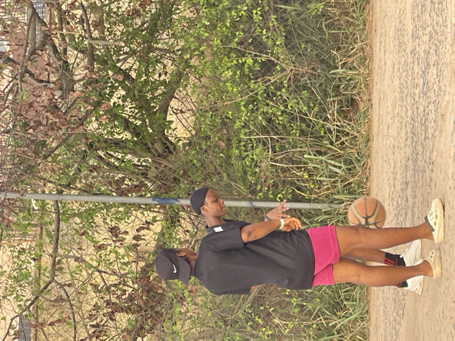 Two people stand on a concrete surface near greenery, including a tree. One person, wearing a black t-shirt, pink shorts, and light-colored sneakers, bounces a basketball. Another person stands close by wearing a black cap, a black t-shirt, and dark socks. A long, thin, horizontal pole stretches across the image behind the two people.
