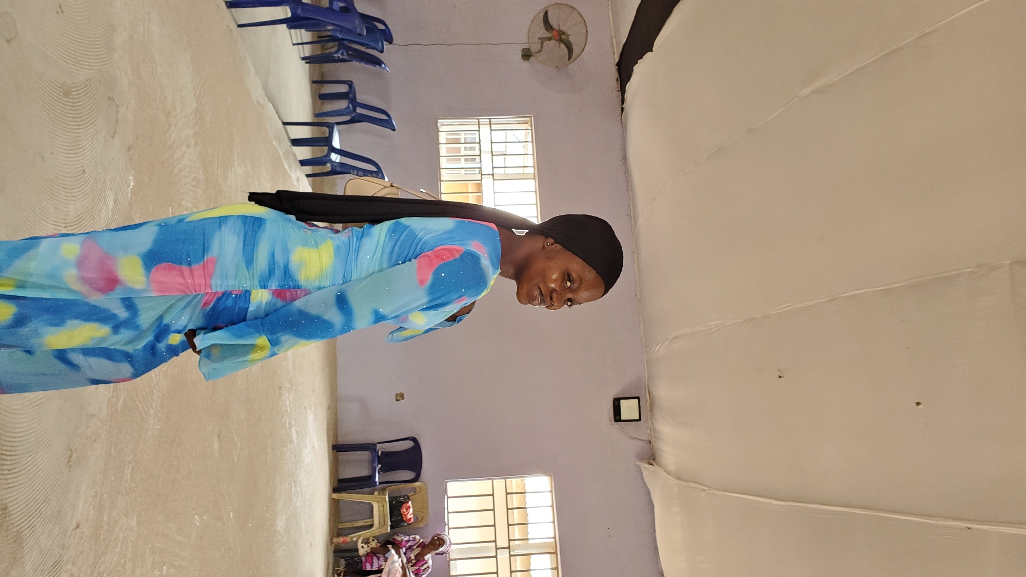 A woman wearing a blue, pink, and yellow tie-dye dress and a black hijab stands in a room with a light purple wall and a white tented ceiling. She is looking over her shoulder at the camera with a smile. To her left, a wall-mounted fan is visible. In the background, there are rows of blue chairs, two windows with barred panes, and a woman sitting in a chair to the right. The floor is a light beige color with circular patterns. The overall setting appears to be an indoor gathering space in Jalingo, Nigeria.