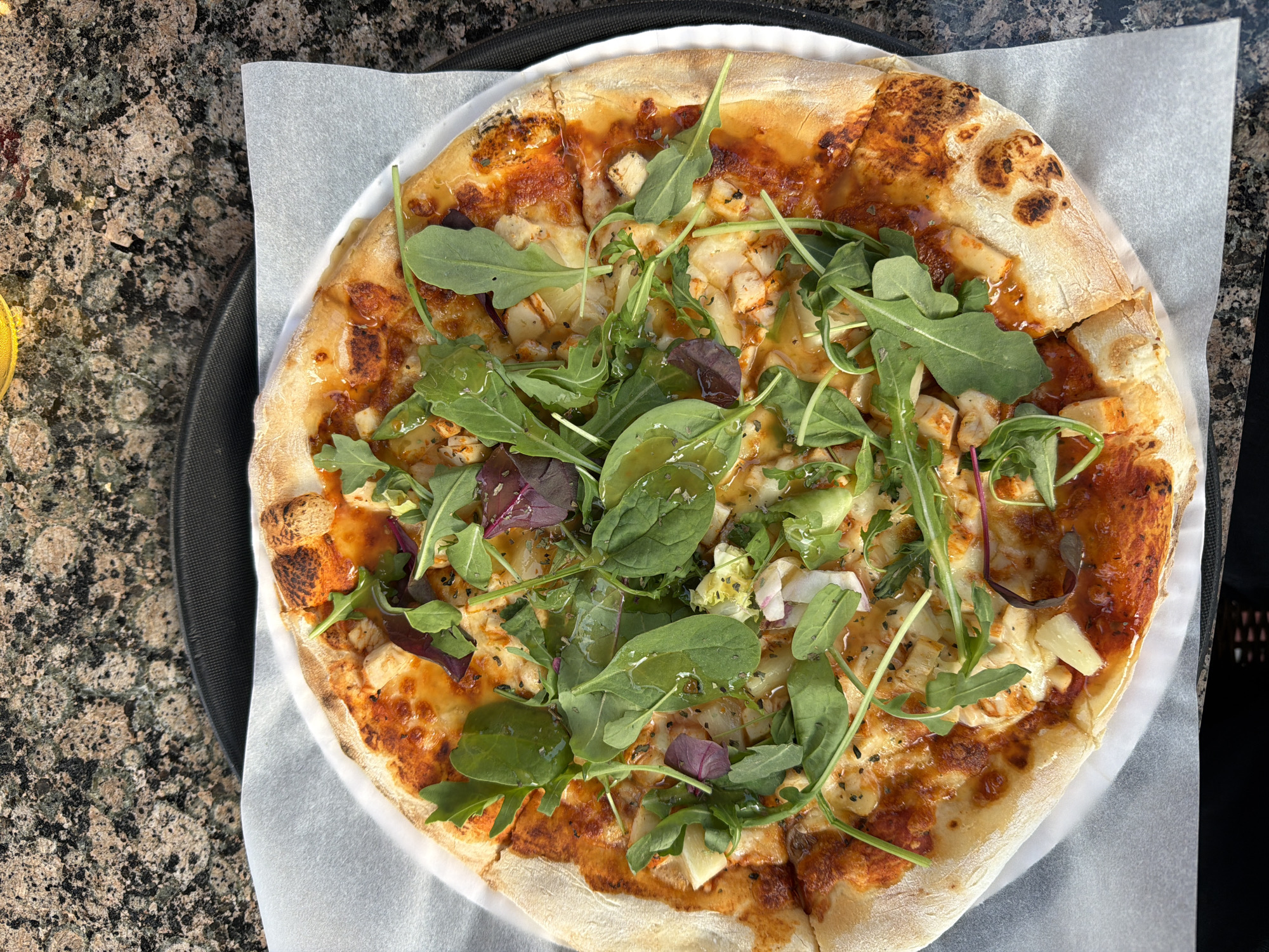 An overhead shot shows a pizza served on a white paper liner on a black tray. The pizza has a golden-brown crust and is topped with red sauce, melted cheese, and pieces of chicken. A generous portion of arugula and some red leaf lettuce are scattered on top of the pizza, glistening with dressing. The pizza is cut into slices and appears to be freshly prepared. The background is a mottled brown and black speckled countertop, possibly granite. The image is sharp and well-lit, highlighting the textures and colors of the food. The scene is likely from an indoor setting, given the lighting and countertop surface. This image was taken in Rūdiškės, Lithuania.