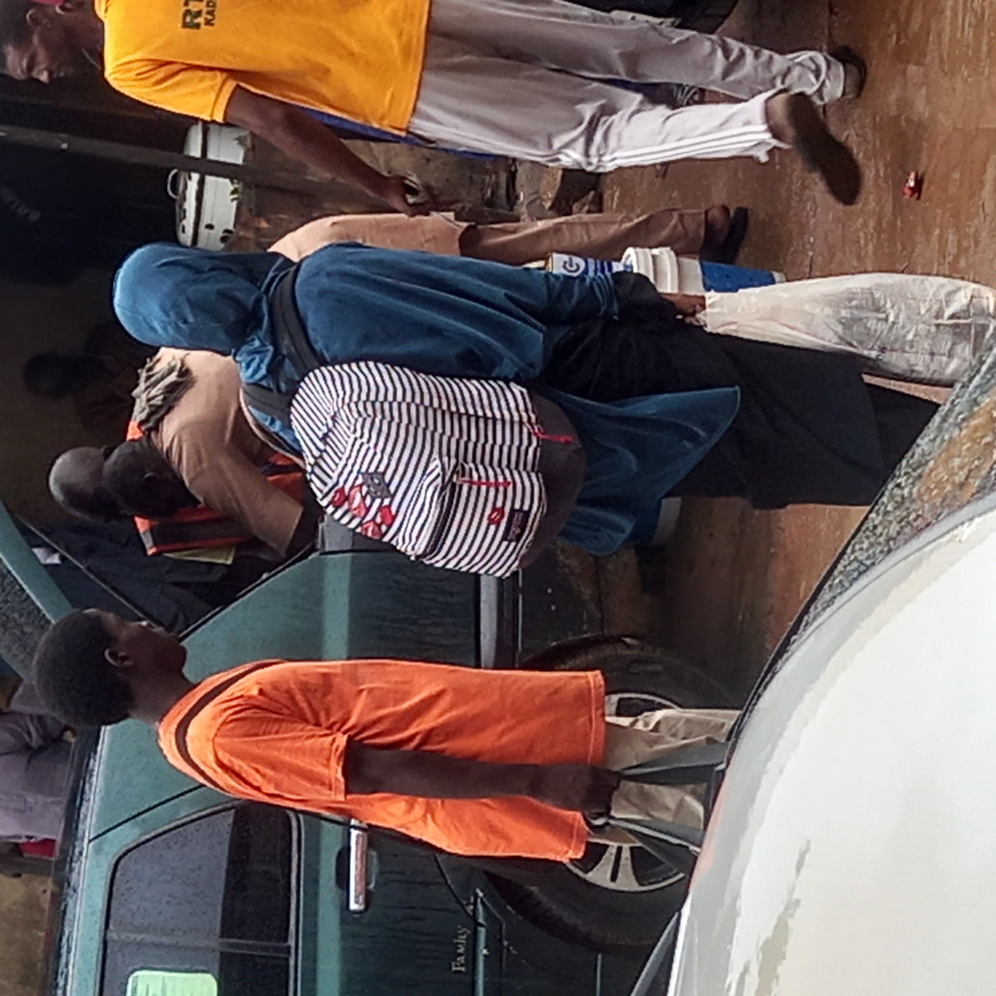 The image, taken in what appears to be a public outdoor space in Dadi Riba, Nigeria, features several individuals engaged in different activities. One man wears a yellow t-shirt with "RT KADE" printed on it and white pants with stripes down the sides. He is walking away from the camera. Another individual in a blue hooded top and black bottoms is wearing a black and white striped backpack. A man in an orange t-shirt stands beside a green vehicle. Other individuals are also visible, their clothing varying in color. The scene is crowded, with a white vehicle partially visible in the foreground.