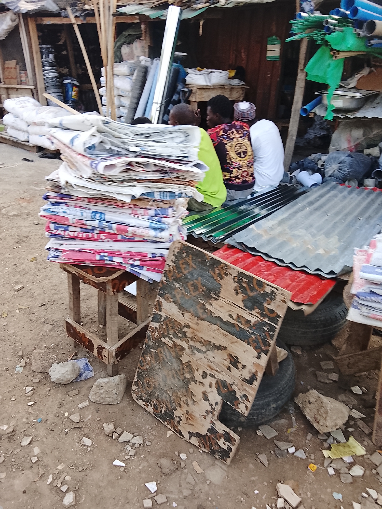 The image depicts a cluttered outdoor scene in Abuja, Nigeria. Three individuals are seated in the background. Two men are wearing a white cap and a gold-patterned shirt respectively. A stack of woven plastic bags sits on a worn, wooden stool in the foreground. Adjacent to the bags are rusted corrugated metal sheets and a weathered piece of plywood leaning against used tires. The ground is littered with debris and rocks. A wooden shack structure with various materials, including plastic pipes and bags of cement, forms the backdrop.