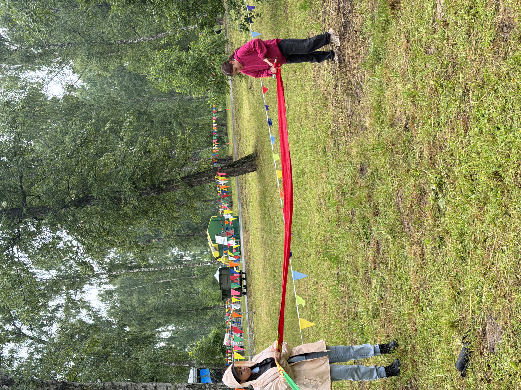 The image depicts two young girls holding a red ribbon stretched horizontally between them in an outdoor park-like setting. The girl on the left is wearing a light-colored hooded jacket and blue jeans. The girl on the right is wearing a bright pink hooded sweatshirt and dark pants. They are standing on a grassy area with trees in the background. There are colorful triangular flags strung along a wire in the middle ground, and in the far background, there are tents and what appears to be a trailer. The ground is covered in grass with some patches of mud. The sky is overcast. The overall impression is that of an outdoor event or competition.
