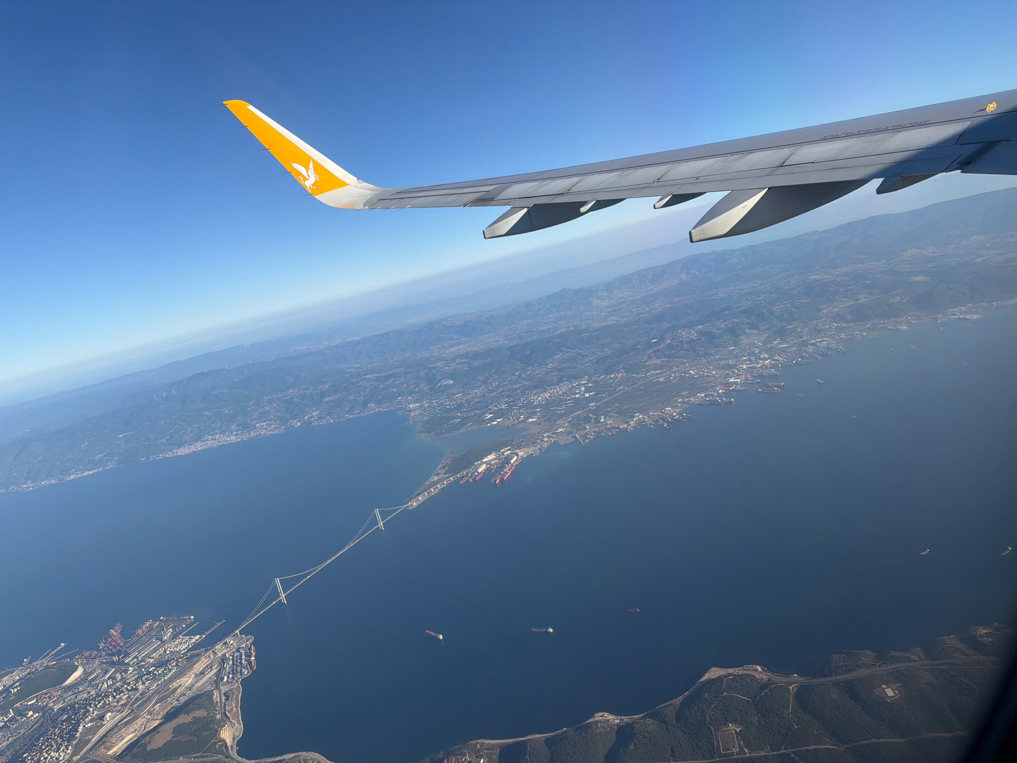 An aerial photograph, taken from an airplane, shows a bright blue sky and a vast expanse of dark blue water. The wing of an airplane, emblazoned with a yellow Pegasus logo, dominates the upper right portion of the image. Below the wing, a long, suspended bridge stretches across the water, connecting two landmasses. The bridge appears to be the 1915 Çanakkale Bridge, located in the Lapseki district of Çanakkale Province, Turkey, near the city of Gebze. The shoreline is lined with buildings and industrial structures, including a port with numerous ships docked. The water is dotted with several cargo ships, some appearing as small specks in the distance. The land beyond the shoreline is hilly and covered in vegetation. The overall scene captures a majestic view of a modern engineering marvel juxtaposed with natural landscapes and maritime activity.