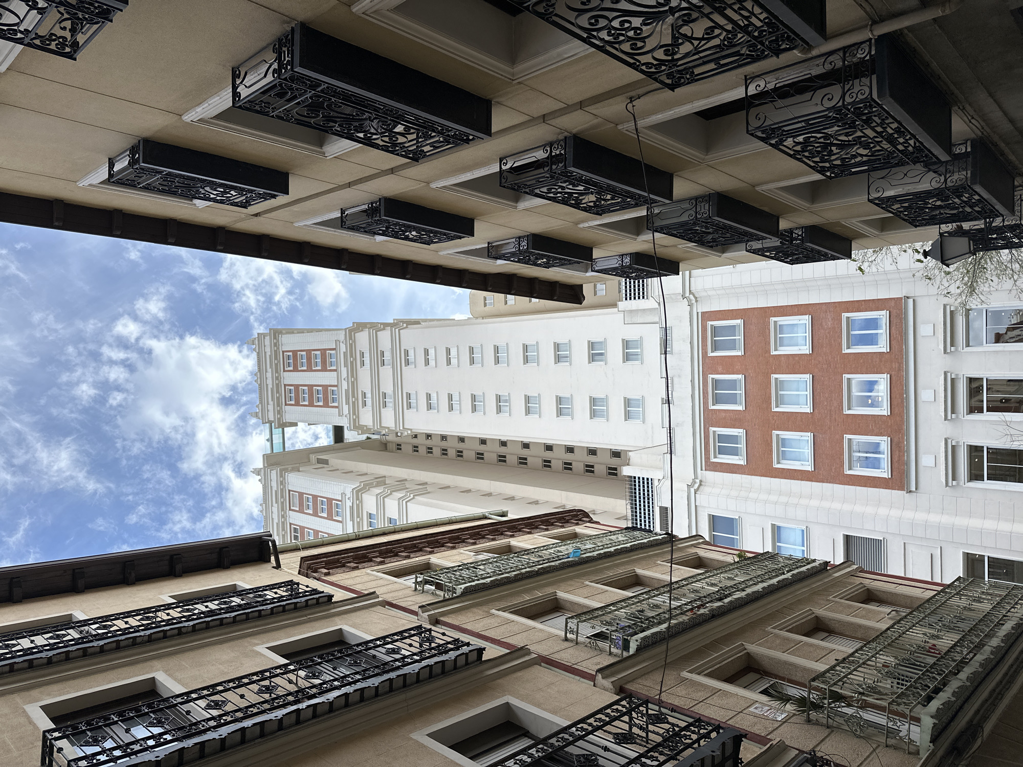 The image is taken looking upward from a narrow street canyon in Madrid, Spain, capturing the architecture and a sliver of the sky. Buildings line both sides of the street. The architectural styles are diverse, with varying facade treatments.
On the left, the building features light brown walls with recessed windows, each adorned with ornate black wrought-iron balconies.
On the right, the buildings present a mix of white and red brick facades. Windows are arranged in regular patterns, and the architectural details are more classical.
In the center, the Telefónica Building, a high-rise structure, rises above the surrounding buildings. It has a mostly white facade with symmetrical window placement. A narrow strip of blue sky and white clouds is visible between the buildings.