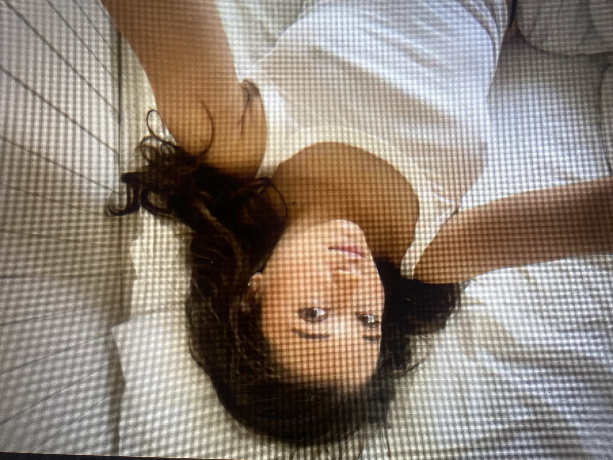 A young woman with dark, curly hair is lying on a white bed with white bedding and pillows. She is looking up at the camera, with her head tilted to the side. She is wearing a white tank top, and her right arm is extended towards the camera. The background is a white wall with vertical wooden planks. The lighting is soft and natural, and the overall mood of the image is relaxed and intimate.