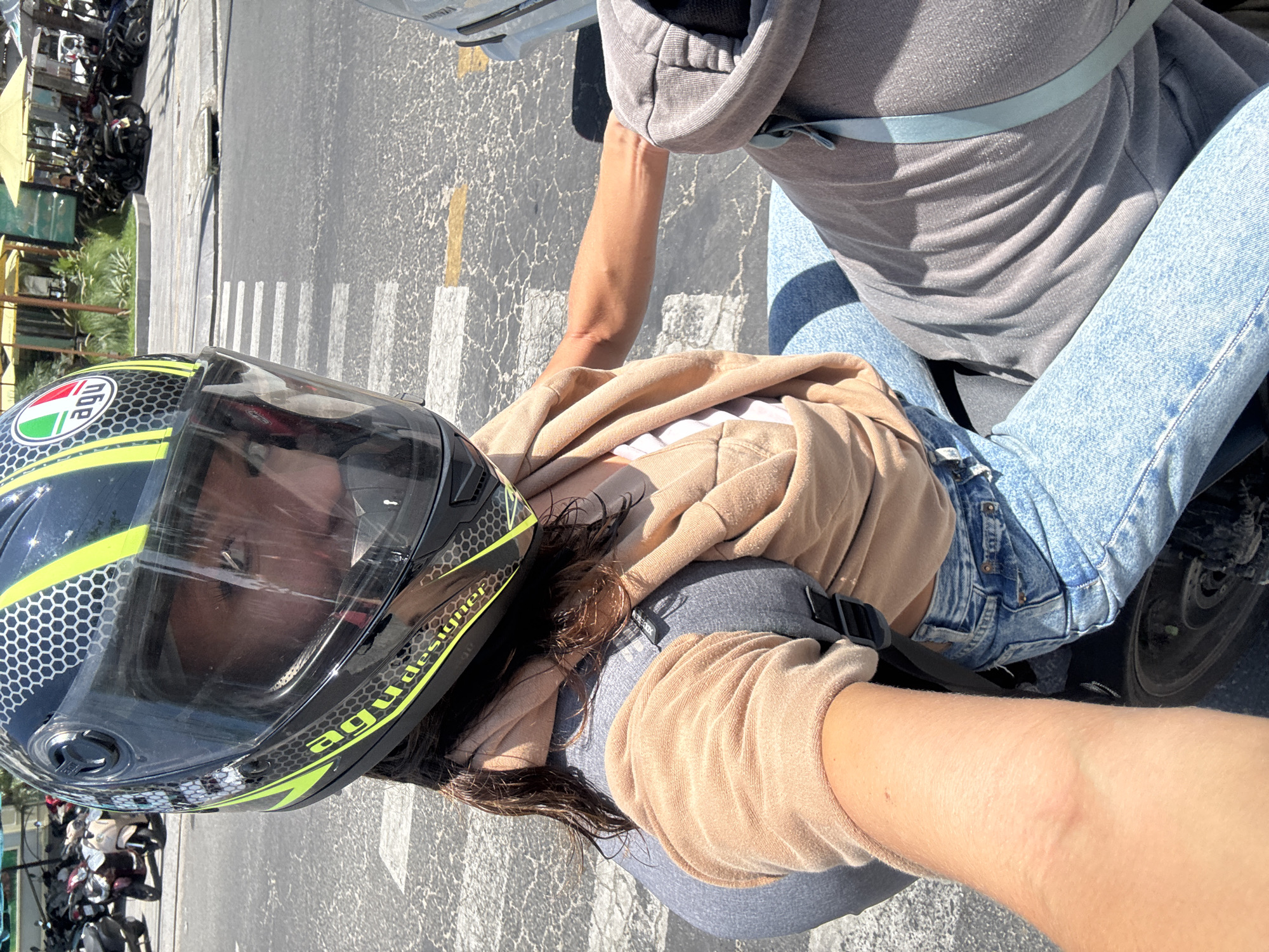 A first-person view from a person on a motorcycle in Đà Nẵng, Vietnam. The person is wearing a helmet with a clear face shield, a tan sweater, and holding onto the motorcycle with their right hand, which is wearing a glove. The road is visible, with a crosswalk painted on the asphalt. Another person is sitting in front of the first person, wearing blue jeans and a gray sweatshirt, a backpack with blue stripes. The background shows a row of parked motorcycles and scooters along the edge of the road. Buildings and trees are also visible in the background.