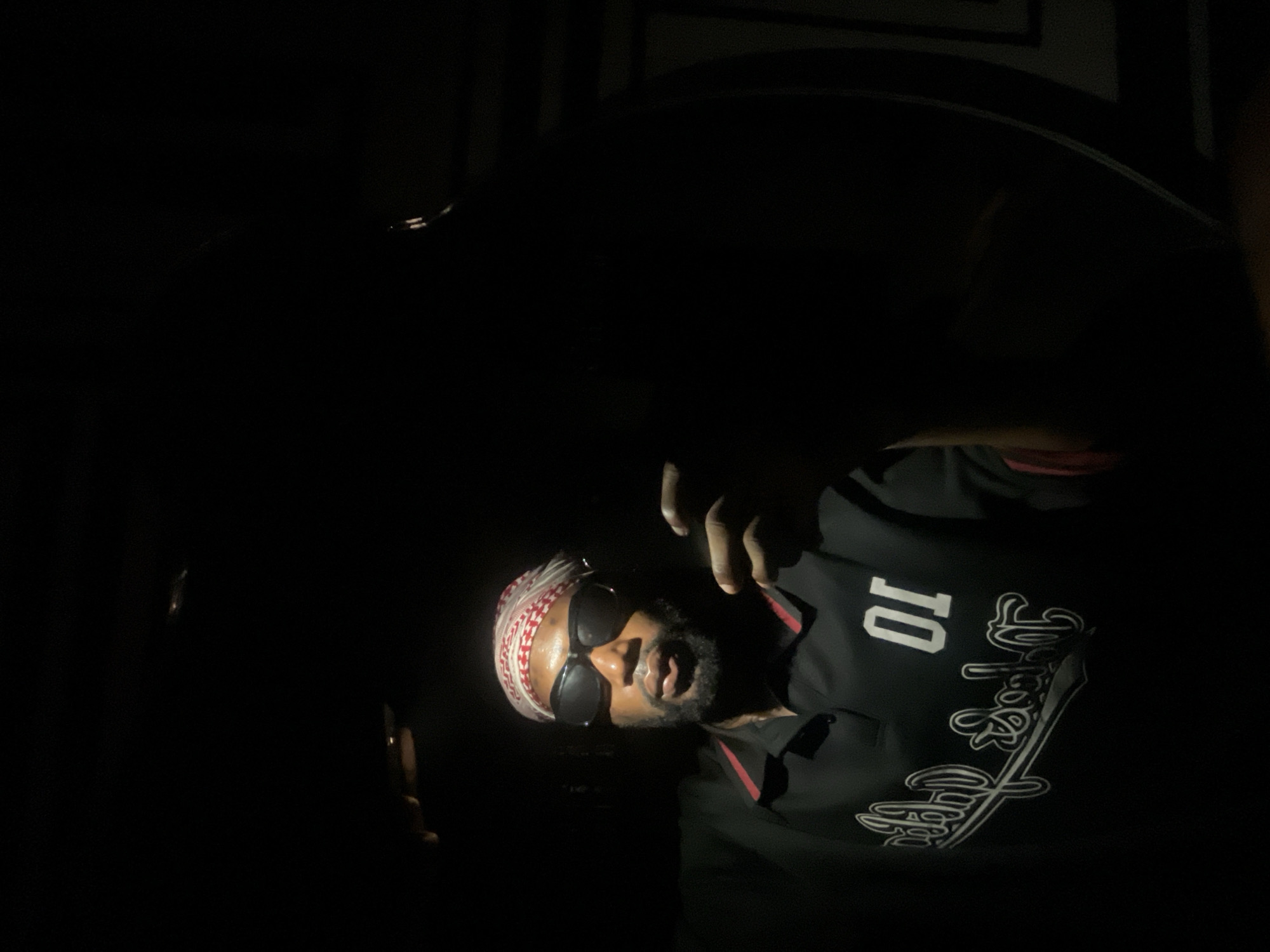A dimly lit scene focuses on a man wearing sunglasses and a patterned headscarf. He is wearing a dark-colored polo shirt with a number "10" and stylized writing on the front. He is partially illuminated by a light source that reveals part of his face and upper body. The background is dark and indistinct. There is not enough information to place this person, or the scene, in Lokoja, Nigeria.