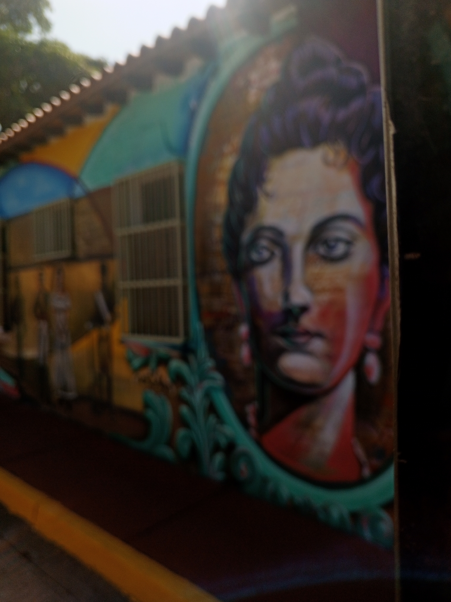 The image displays a vibrant street mural in La Asunción, Venezuela, painted on the side of a building. The central feature of the mural is a stylized portrait of a woman, depicted from the shoulders up, framed by an elaborate, ornate oval border. The woman has dark, curly hair styled in an updo, light-colored eyes, and her face is rendered with a dynamic blend of skin tones, red, and orange accents, suggesting a bold artistic style. She appears to be wearing earrings.
To the left of the portrait, the mural continues to show a section of a building with several rectangular windows, some equipped with vertical bars. Below these windows, several figures are vaguely visible, appearing as blurry human forms, possibly representing people walking or standing on a street. One figure in the foreground on the left wears light-colored attire.
The mural utilizes a rich and colorful palette, incorporating bright blues, greens, yellows, and oranges in the background elements, contrasting with the warmer tones of the woman's face and the brick-like texture behind her. A corrugated or tiled roofline is visible along the top of the building, illuminated by what appears to be bright sunlight, suggesting the scene takes place during the daytime. A yellow line, characteristic of a street curb, runs along the bottom left edge of the frame. The overall image has a somewhat soft or out-of-focus quality, particularly noticeable on the left side and the lower portions, adding to its artistic and atmospheric feel. No discernible text is visible within the image.