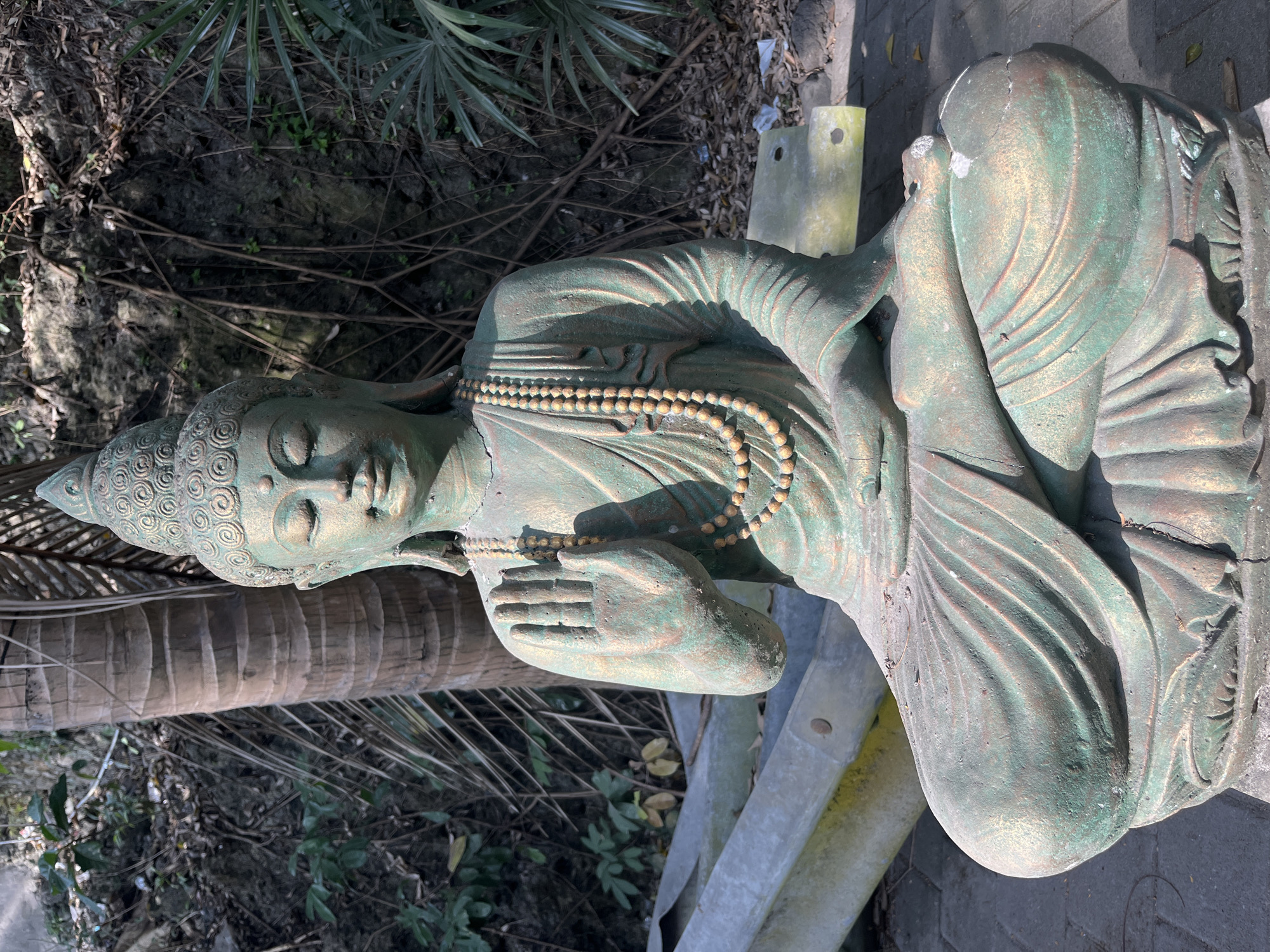In Kuta Selatan, Indonesia, a weathered statue of the Buddha sits in a meditative pose on a paved surface. The statue, predominantly green with hints of gold, is adorned with prayer beads around its neck. Its eyes are closed, and one hand is raised in a gesture of blessing or protection. The background features a rocky incline with sparse vegetation and a palm tree trunk to the left. Discarded materials, including what appears to be part of a metal structure, lie in the foreground and to the sides. The lighting suggests it is daytime, with shadows indicating the sun's position.