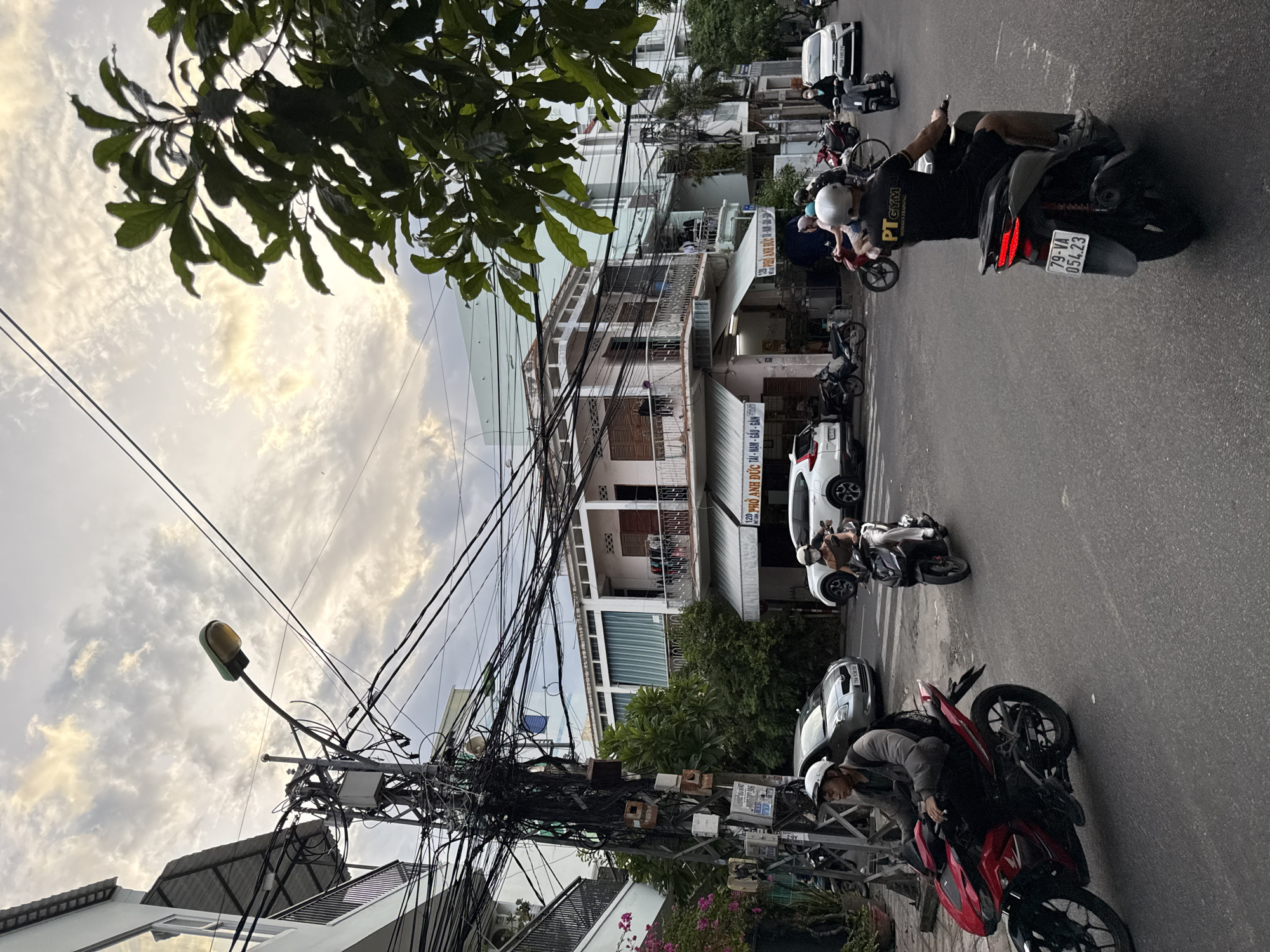The image depicts a street scene in Nha Trang, Vietnam, likely during daylight hours given the illumination. The foreground is dominated by a paved road with several motorbikes in motion. On the left, a person wearing a gray jacket and a white helmet rides a red motorbike. In the middle ground, another person on a dark-colored motorbike is visible, with the text "PT GYM" on their black shirt. Further back, other motorbikes and a silver car are present. The background features multi-story buildings with balconies and awnings, characteristic of urban Vietnamese architecture. A prominent utility pole laden with a dense network of electrical wires stands on the left side of the street. Overhead, a streetlamp with a green fixture is visible against a cloudy sky with patches of sunlight. Lush green foliage from trees overhangs parts of the scene. The overall impression is one of a bustling urban environment with a constant flow of motorbike traffic.