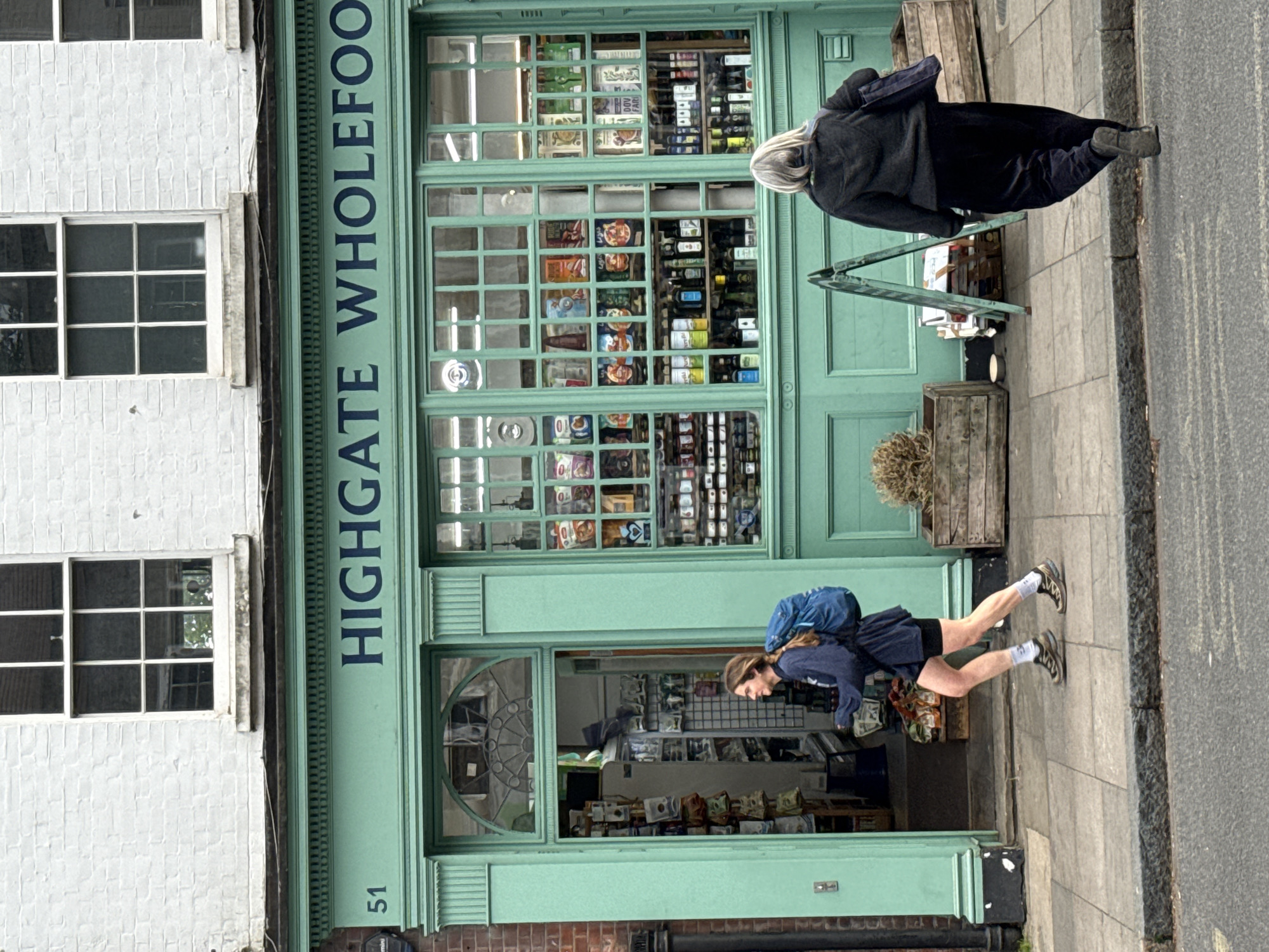 The image shows the exterior of Highgate Wholefoods, located in Highgate, London, United Kingdom. The building facade is painted a light green color, with the shop name "HIGHGATE WHOLEFOODS" in dark green lettering above the shopfront. The number "51" is visible beneath the shopfront on the left.
The shopfront features a large display window with numerous small panes, filled with various grocery items. The entrance has a decorative arched window above the door. A white brick building is adjacent to the shop on the left, with visible windows.
Two pedestrians are present: one older individual with white hair wearing a dark coat and black bottoms is walking away from the shop on the right. Another younger individual with a backpack, wearing a dark top and shorts, with white socks, is walking into the shop. A small triangular sign and some potted plants are placed outside the shop on the sidewalk. The sidewalk is grey and a road is visible on the right edge of the image.