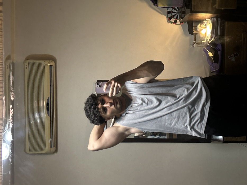 A person stands in front of a mirror, taking a selfie. They are wearing a gray tank top and dark bottoms, with dark curly hair. To the left, an air conditioner unit is mounted on the wall. Behind the person, a dark wooden cabinet holds various items, including a dartboard and what appears to be a lamp or light fixture. The room has a neutral-colored wall, with the mirror positioned vertically.