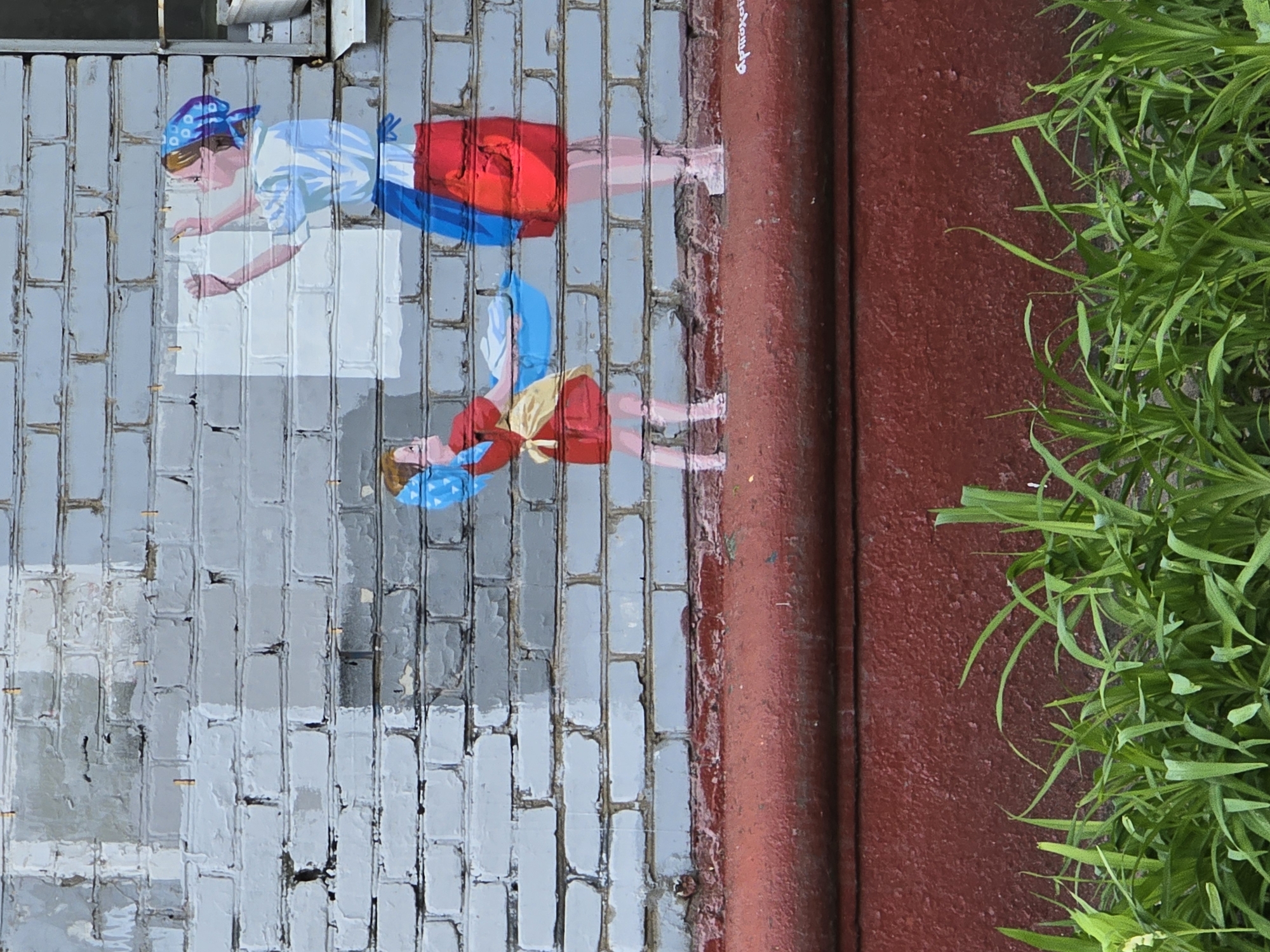 The image depicts a mural on a gray brick wall, featuring two figures. The upper figure is a woman wearing a white shirt, red skirt, and blue headscarf, reaching out with her hands as if interacting with a blank white rectangle on the wall. Below her, a similar but smaller figure, possibly a child, wears a red dress, yellow sash, and blue headscarf, also reaching out. To the right of the brick wall is a reddish-brown painted surface and green vegetation. The location is Khimki, Russia.