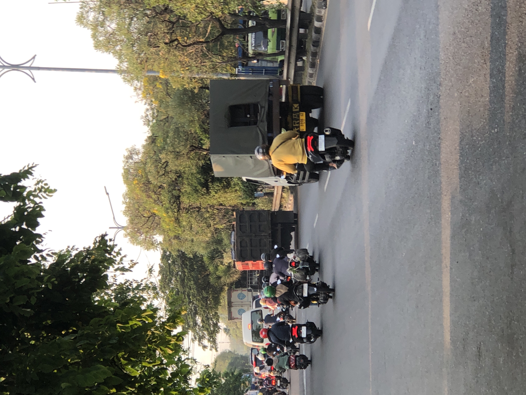 This image captures a bustling street scene in Kludan, Indonesia, likely during daylight hours given the bright illumination and clear sky. The foreground is dominated by a paved road with lane markings, where numerous motorcycles are in motion. Most of the riders are wearing helmets, and some are accompanied by passengers. The motorcycles are of various colors, and several have cargo strapped to the back.
In the middle ground, larger vehicles are present, including a white van and several trucks. One truck, dark grey with a canvas covering, is prominently positioned. Another truck, red and black, is also visible. Behind these, a bright green truck can be seen further down the road. Trees line the sides of the road, some with yellow flowers, contributing to a tropical or sub-tropical environment. A tall lamppost is visible on the right side of the image, suggesting an urban or suburban setting. The overall impression is one of typical traffic flow in a busy Indonesian city.