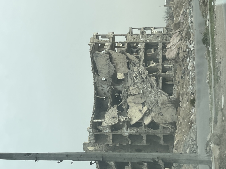 The image depicts a severely damaged urban landscape within Joubar Municipality, Syria. The primary subject is a multi-story concrete building, likely a former residential or commercial structure, exhibiting catastrophic damage. The building's facade is largely destroyed, revealing interior floor slabs, exposed rebar, and extensively shattered walls. Significant portions of the structure have collapsed inwards, creating large piles of concrete rubble and debris. Numerous circular impact marks, consistent with shrapnel or bullet damage, are visible on the remaining concrete surfaces.
In the immediate foreground on the left, a damaged utility pole, possibly made of concrete, stands vertically with multiple visible perforations and fractured sections. A paved road or street traverses the lower part of the frame. The ground between the road and the damaged building is covered in extensive debris, including fragmented concrete, twisted metal, and shattered masonry. Sparse, bare vegetation is visible amidst the rubble. The sky above is overcast and uniformly light grey. No individuals or active movements are present in the scene, which conveys a desolate and war-affected environment.