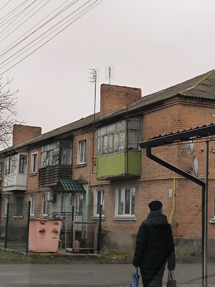 The image presents an overcast daytime scene of a street in Illintsi, Ukraine, featuring a multi-story brick residential building. The building, likely an apartment block, has a weathered grey roof and several enclosed balconies/loggias, each with a distinct appearance; one on the right is painted bright green, another in the middle has dark corrugated siding, and one on the left is white. Multiple windows are visible across the facade. Two Yagi-style television antennas are mounted on the rooftop, above brick chimney structures.
In the foreground, a person is seen from behind, walking away from the viewer on a paved surface next to the building. The individual is dressed in a dark, possibly padded, winter coat and a dark hat, carrying a blue-patterned bag in one hand and a dark bag in the other.
Notable details include overhead utility lines against the grey sky on the left. An entrance to the building features a small green arched awning and a dark metal gate. In front of this, there is a large, faded pinkish-orange dumpster-style trash bin and a metal grate enclosure, likely for refuse collection. Green metal posts mark a boundary. On the brick wall to the right, the number "10" is affixed, possibly a building or entrance number. Two white satellite dishes are visible on the building's right side, below a black rain gutter system. The scene depicts a quiet, everyday moment in a typical Eastern European residential setting under a cloudy sky.