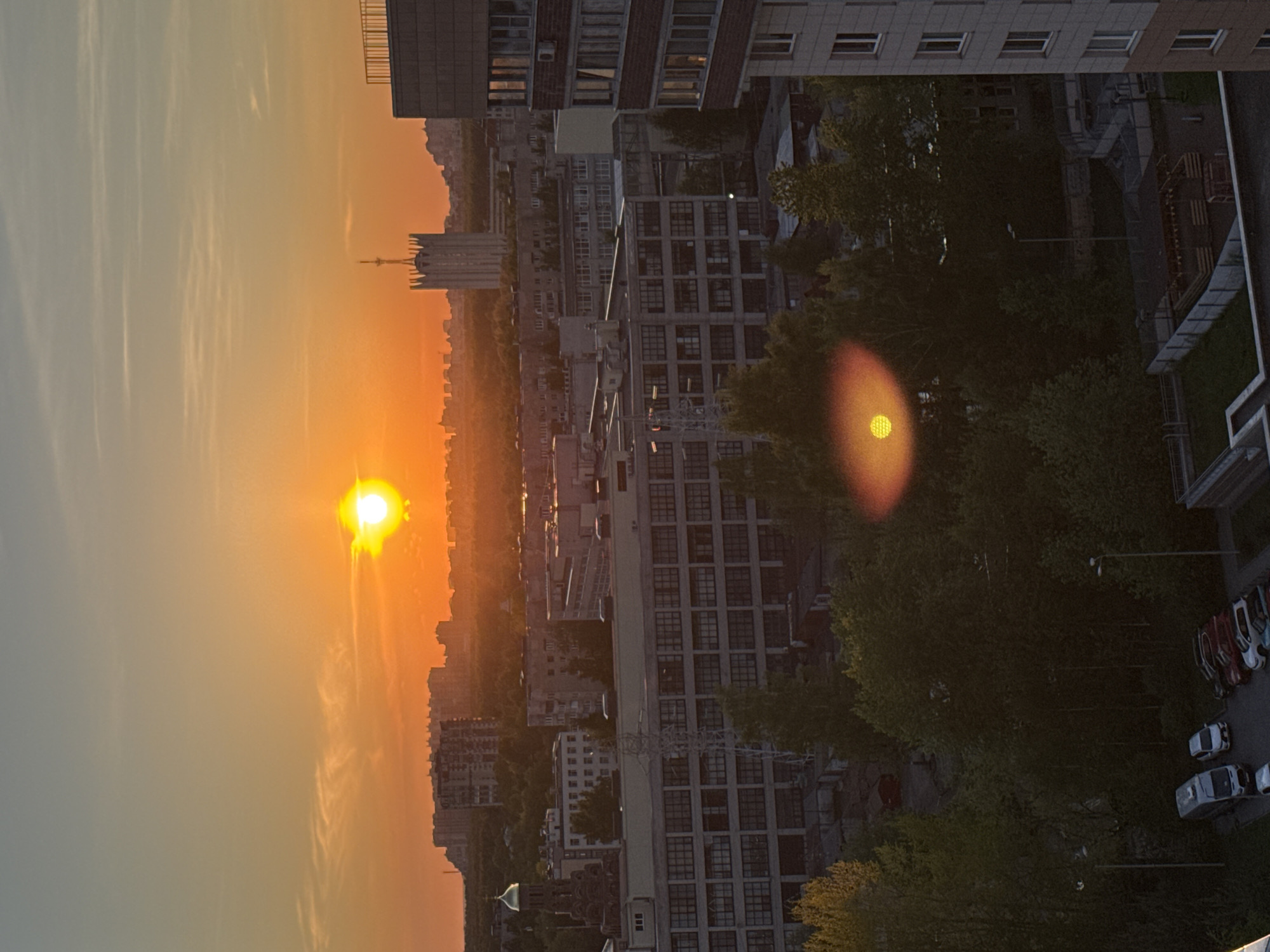 The image depicts a cityscape at sunset, possibly in Saint Petersburg, Russia, considering the architectural style of the buildings and the general urban layout. The sun is low on the horizon, casting a golden light over the city, with lens flare visible. Multiple high-rise residential and commercial buildings are evident, forming a dense urban area. The presence of trees suggests parks or tree-lined streets within the city. The image also shows parked cars along a street. There are no people visible in the scene.