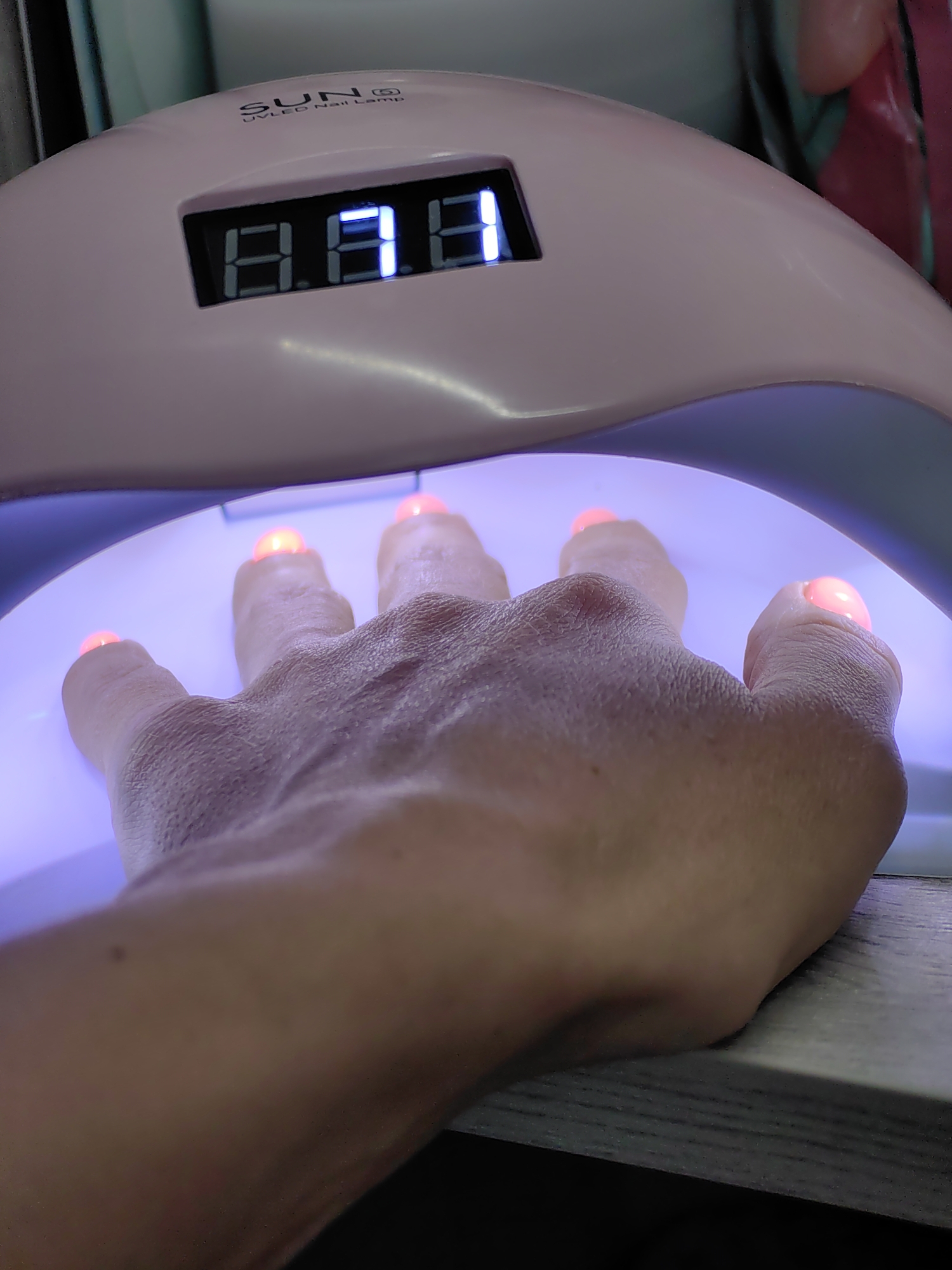 A close-up shot shows a hand with freshly painted nails placed inside a pink UV/LED nail lamp. The lamp has a digital display showing "8.8.8". The nails on the fingers are illuminated with a pinkish-orange glow, indicating they are being cured by the lamp. The background is out of focus, but suggests an indoor setting. The image is taken from a slightly elevated angle, looking down at the hand. The scene is indoors in a room.