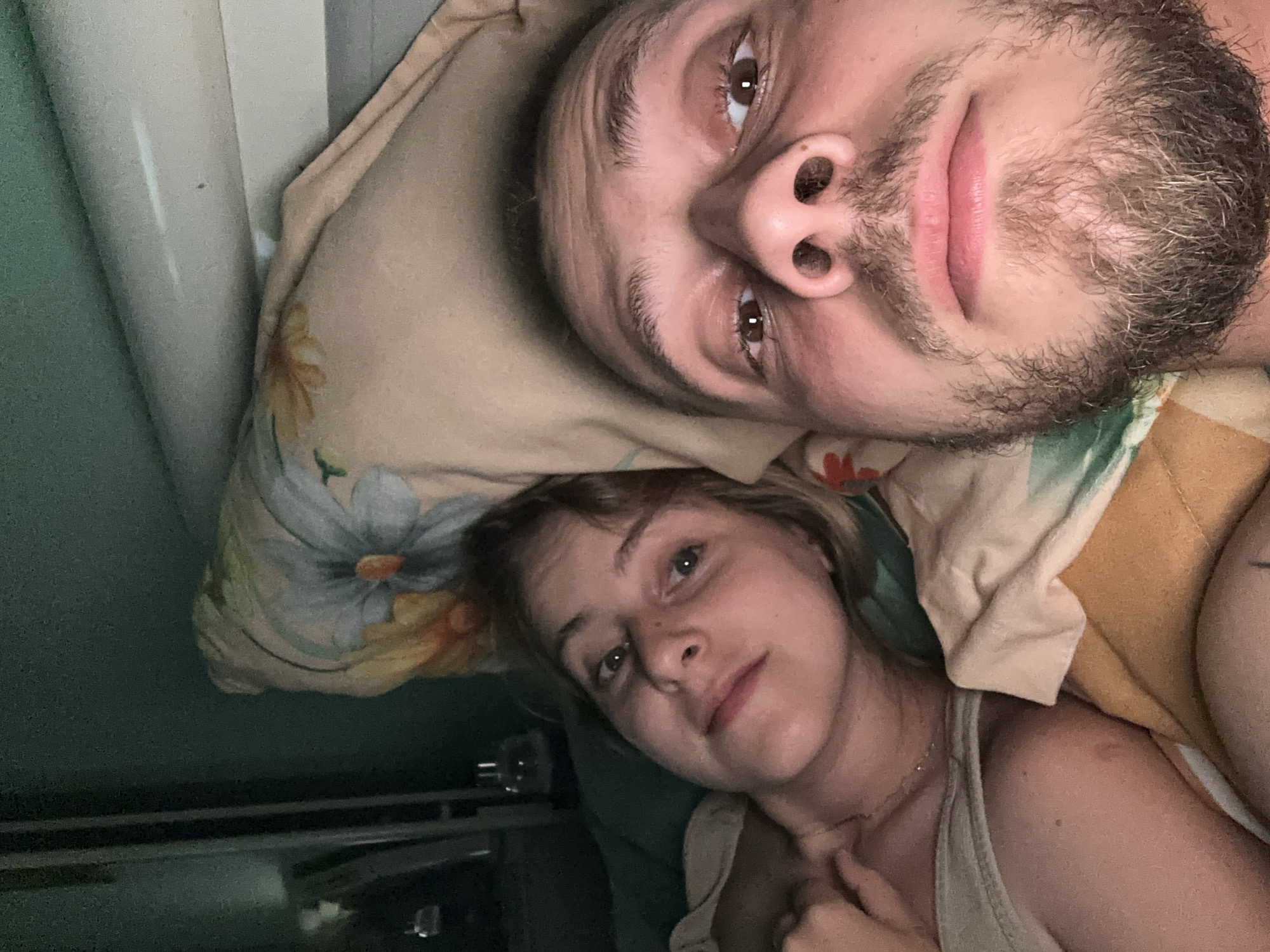 Two people are lying in bed, facing the camera. The person on the left is a woman with blonde hair, looking towards the camera. The person on the right is a man with a beard, also looking towards the camera. They are under a patterned blanket, and the background appears to be a bedroom. There is a bottle in the background on a shelf. The overall lighting is dim.