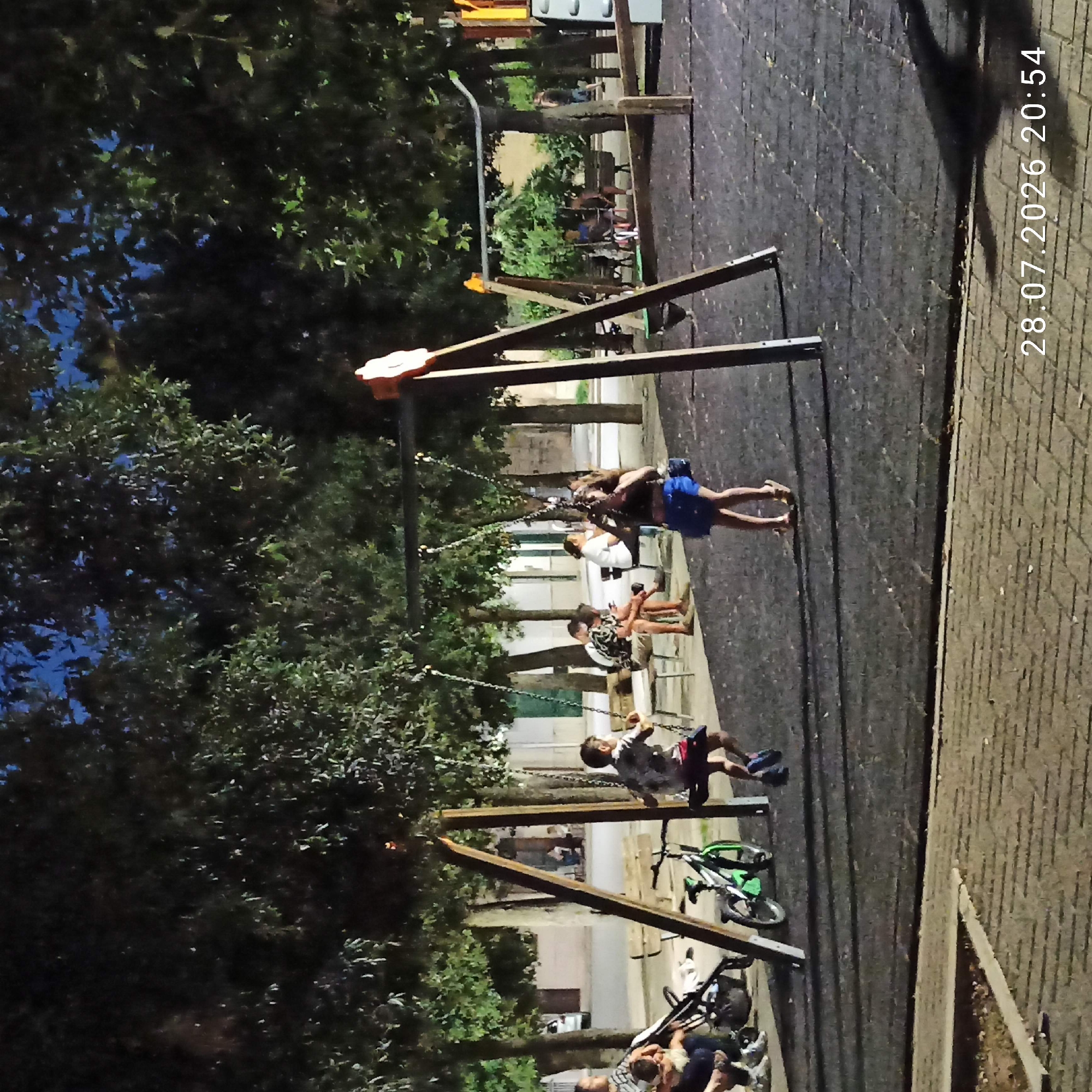 The image depicts a nighttime scene at a playground in Bitonto, Italy. The playground is equipped with swings and a soft, dark rubberized surface. Several people are present, with the most prominent being a young boy on a swing in the foreground, wearing a patterned shirt and dark shorts. To the left, an adult and a child are near a stroller, and a bicycle is parked nearby. In the background, other individuals are seated on benches and standing, engaged in various activities. The playground is surrounded by trees, and buildings are visible in the distance, illuminated by streetlights and ambient light, suggesting an urban or semi-urban setting. The date and time stamp "28.07.2026 20:54" is visible in the bottom right corner of the image.