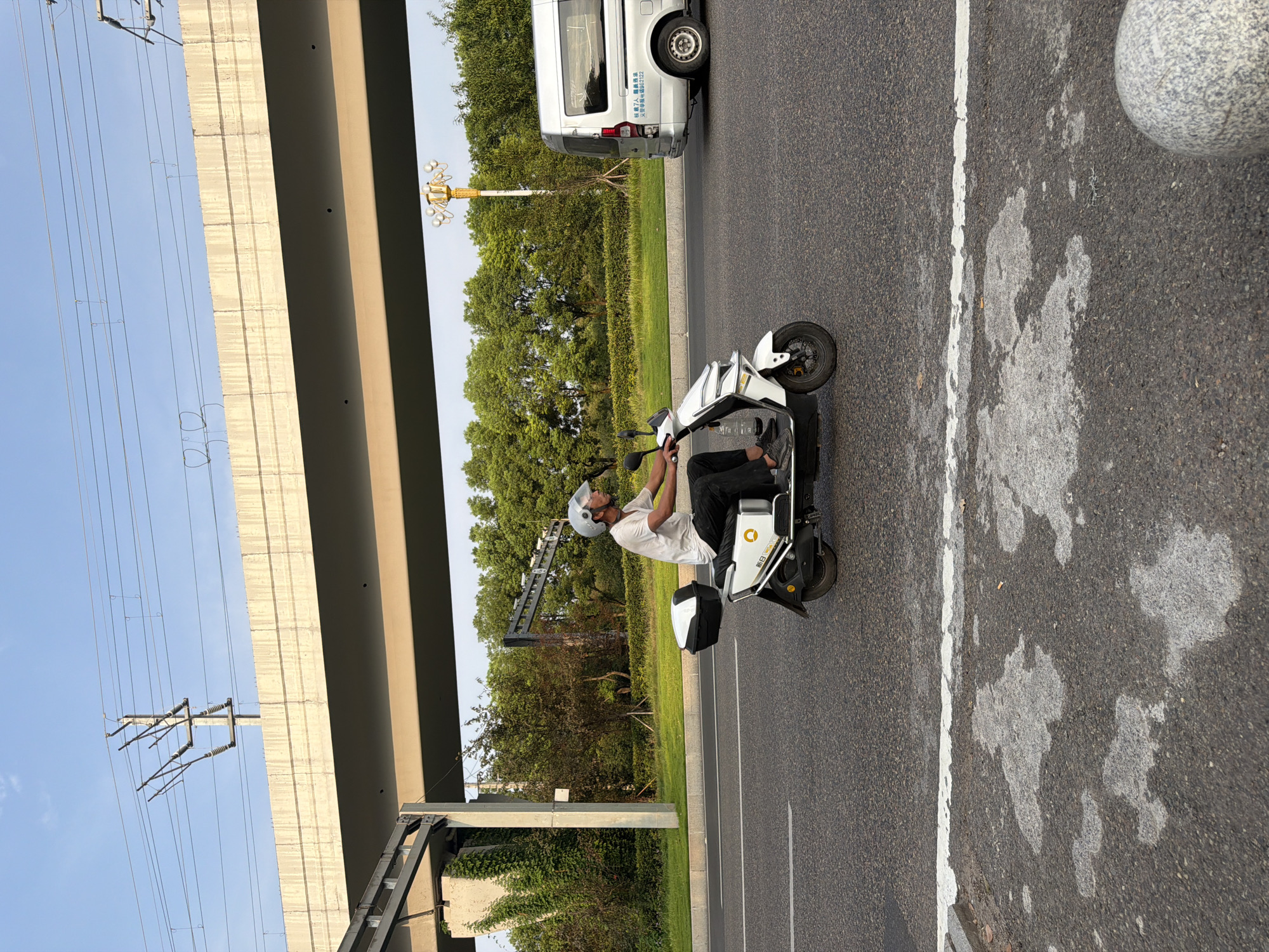 A man wearing a white helmet and a white t-shirt rides a white electric scooter with a black storage box on the back, traveling on a paved road. He is wearing dark pants and dark shoes. The scooter has a white logo with yellow lettering on the side. In the background, there is a multi-lane elevated concrete structure, likely an overpass or bridge, with overhead power lines. Beyond that, a dense line of green trees forms a backdrop. To the right of the scooter, a silver minivan is visible on the same road. The road surface is asphalt with white lane markings. The scene appears to be in an urban or suburban environment, possibly in the city of Jiangjin, China, given the context. The sky is a clear blue with some light clouds. The lighting suggests it is daytime, likely late afternoon due to the angle of the shadows.