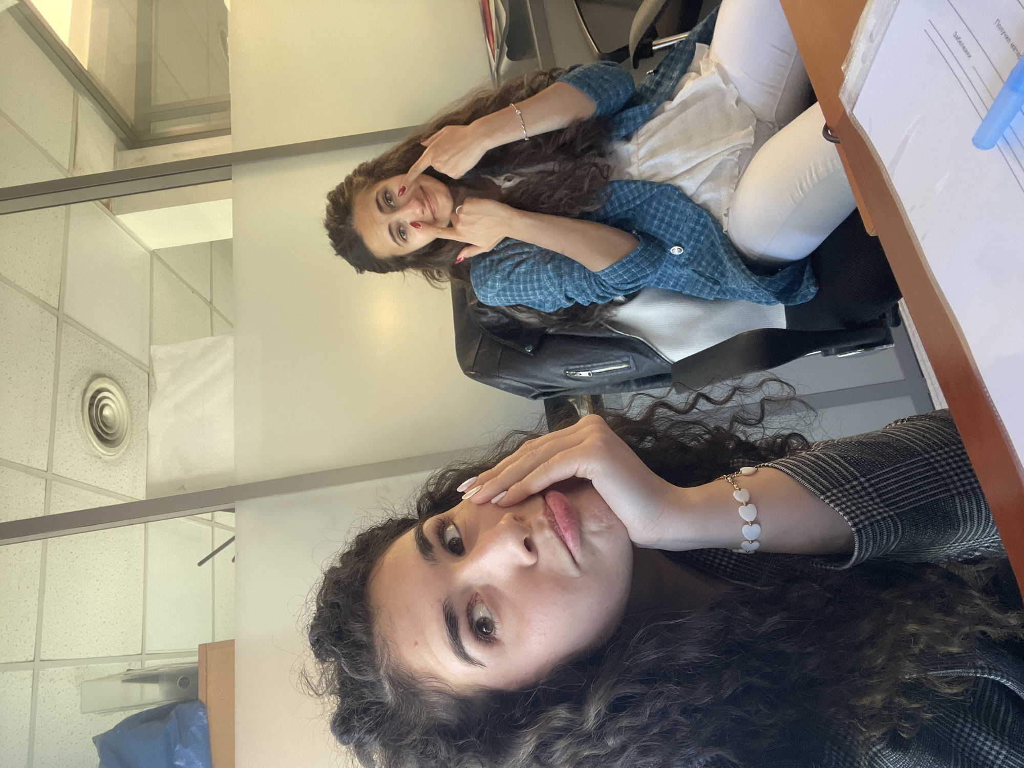 The image shows two women in what appears to be an office environment. The woman in the foreground has dark, curly hair and is wearing a patterned blazer. She is resting her face on her hand, looking towards the camera. The second woman, seated in a chair, has long, wavy brown hair and is wearing a blue, checkered shirt and white pants. She is smiling and pointing at her cheeks with her fingers. In the background, there's a wall and office furniture. A file folder is visible on a desk. The location cannot be determined based on the image alone. The city of Sofia, Bulgaria is extraneous information.