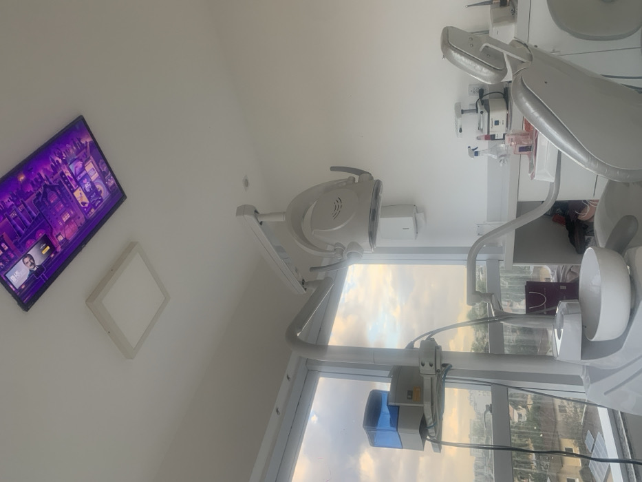 The image depicts an unoccupied, modern dental operatory located in the city of Lauro de Freitas, Brazil. The room features white walls and a white ceiling. Two light fixtures are visible on the ceiling: a square, flush-mounted LED panel and a larger, rectangular display screen exhibiting purple-toned digital art, which includes a partial human profile in the upper left corner.
In the foreground, a grey dental chair is positioned, equipped with a white ceramic spittoon basin and an attached arm supporting a water reservoir with a blue-translucent top. A white dental examination light, designed to illuminate the patient's oral cavity, is suspended overhead and to the right of the chair.
Along the back wall, a white, minimalist countertop holds various dental instruments, including spray bottles, small containers, and a white, table-top appliance, possibly a heating or curing unit. A purple shopping bag is partially visible beneath the counter. A large, floor-to-ceiling window on the left side of the room provides an expansive view of an urban landscape with multiple buildings and trees under a cloudy sky, indicating the operatory is located on an upper floor. No individuals are present in the scene.