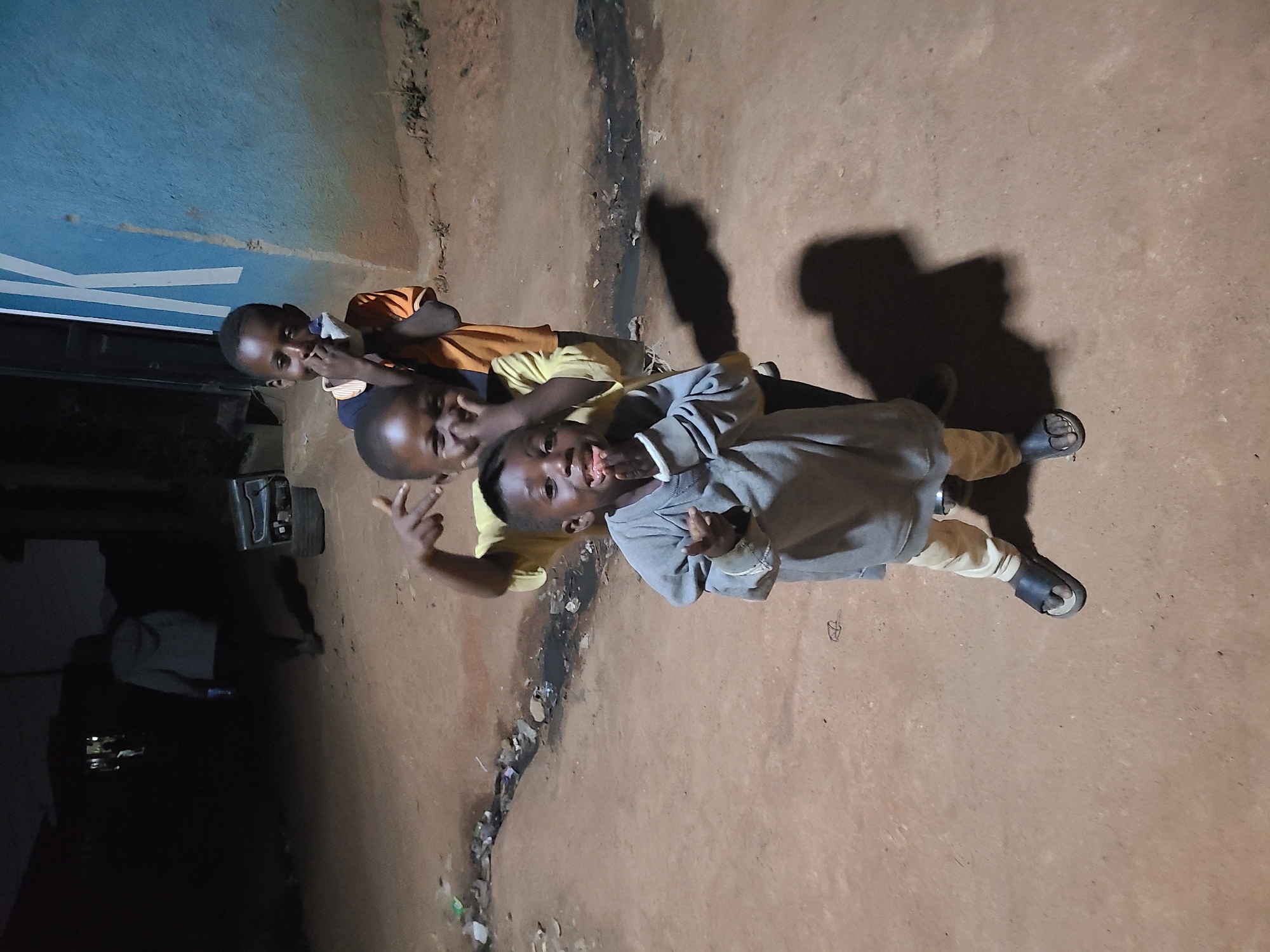 Three young African children are standing in a line, facing the camera. The child in front is wearing a gray sweatshirt and khaki pants, with open-toed sandals, and is making a hand gesture. The second child is wearing a yellow shirt and the third child is wearing an orange shirt. They appear to be outside at night, standing on a dirt or concrete surface next to what seems to be a curb. There is a dark building or structure behind them on the left. The location is in the city of Jos, Nigeria. There is trash or debris scattered along the curb. The children's shadows are cast to their right.
