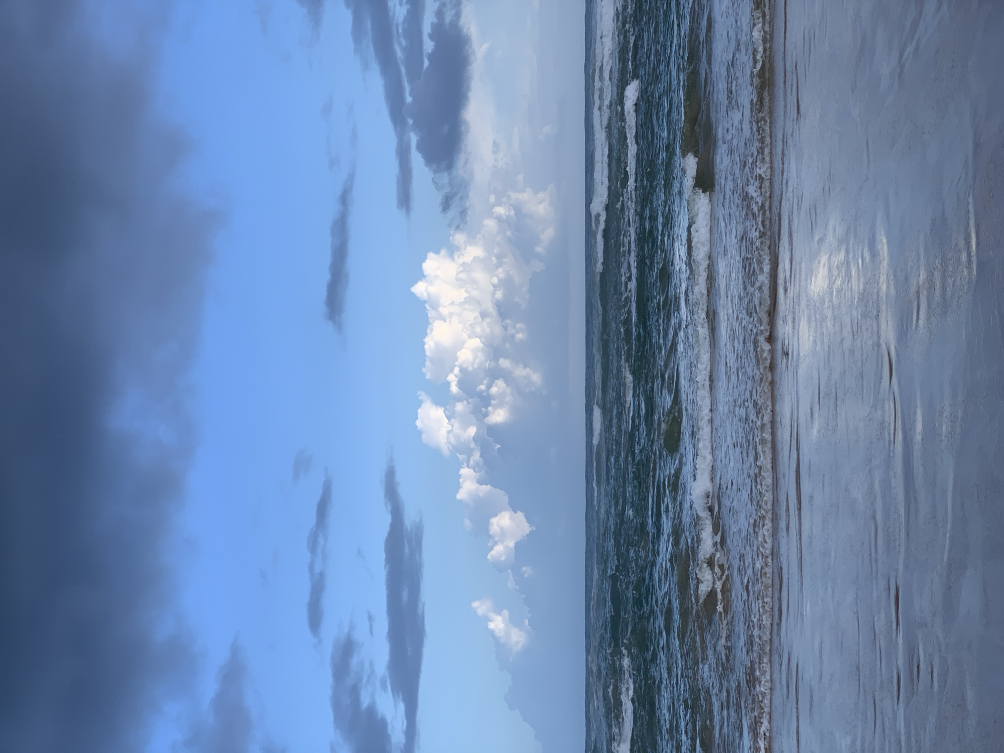 The image shows a view of the beach and ocean in Ahangama, Sri Lanka. The photo is taken in a vertical orientation, with the sky and clouds on the left and the ocean and beach on the right. The sky is a mix of blue and dark gray clouds, with a patch of bright white clouds in the middle. The ocean is a dark teal color with white waves crashing on the shore. The beach is wet and sandy, reflecting the sky and ocean. No people are visible in the image.