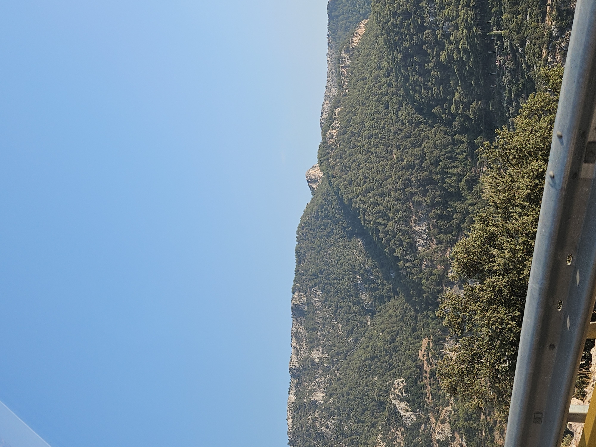 A clear, blue sky occupies the upper half of the image, with a faint gradient indicating the horizon. The lower half of the image features a lush, green mountainside densely covered with pine trees. The rocky, barren terrain of the mountain peaks is visible at the top, with patches of exposed rock interspersed among the vegetation. In the foreground, a grey metal guardrail runs horizontally across the bottom of the frame, separating the viewer from the mountainside. Some of the guardrail posts are visible, along with bolts and other hardware. The lighting suggests it is daytime, with sunlight casting shadows and highlighting the textures of the mountainside.