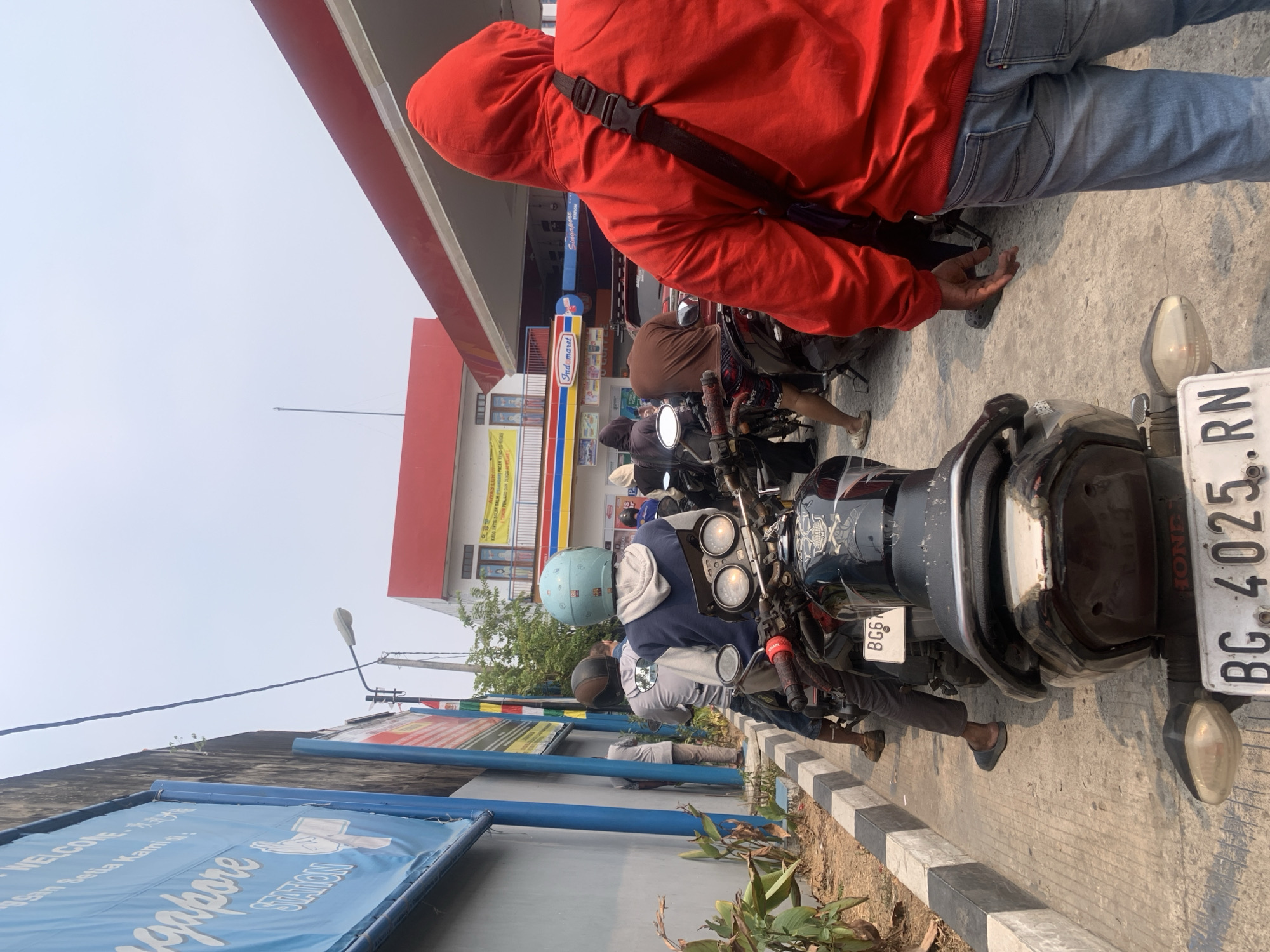 This image depicts a scene at a gas station, specifically Singapore Station, in Palembang, Indonesia. The foreground features the rear of a black motorcycle with a license plate reading "BG. 4025. RN". To the right, a person wearing a red hooded sweatshirt and blue jeans stands facing away from the camera, their left hand resting on their thigh. Behind this individual, other motorcycles and people are visible, suggesting a queue.
In the mid-ground, several motorcycles are lined up, with riders or passengers on them. One motorcycle in the center has a rider wearing a light blue helmet and a dark blue hoodie, and another behind them wearing a gray hoodie. To the left, a person in a gray t-shirt stands by a wall.
The background shows the gas station's structure with a prominent red roof and canopy. A blue banner with the text "Singapore STATION" is visible on the left side of the image. A building with a red roof and white walls, possibly a convenience store (indicated by an "Indomaret" sign), is situated behind the gas pumps. The environment appears to be an outdoor setting with a bright, overcast sky. The ground is paved concrete with a strip of black and white marked curb. Vegetation is present along the wall on the left.