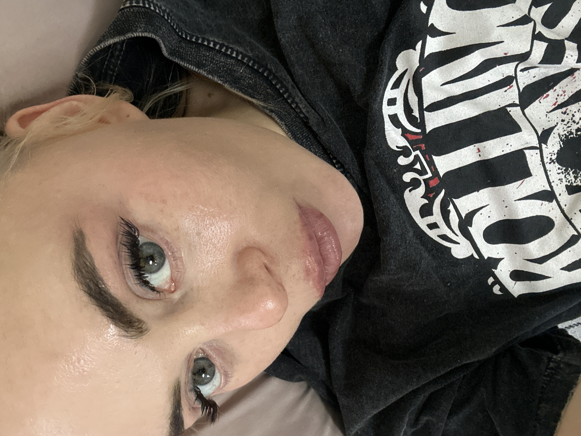 A fair-skinned person with light blonde hair is lying down, as evidenced by the rotated perspective. The person's face is prominent, with focus on their eyes, nose, and lips. They have dark eyebrows and long eyelashes. They are wearing a dark-colored shirt with a white graphic design. The background is a light, solid color, possibly a bed or similar soft surface. There's no information available in the image to associate it with the city of Высокий, Russia.