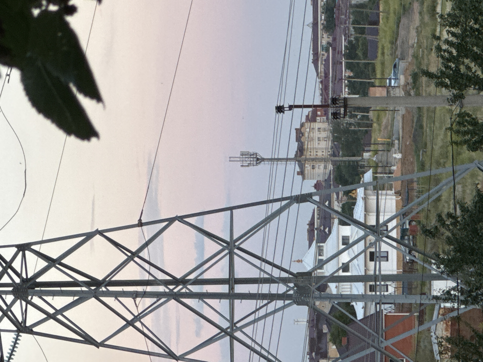 The image depicts a view from behind a large, grey metal electricity pylon. The pylon's lattice structure dominates the foreground, with diagonal beams forming a complex geometric pattern. In the background, a hazy sky transitions from pale pink to a light blue, suggesting either dawn or dusk.
Beyond the pylon, a streetscape unfolds. Several multi-story buildings with light-colored facades and dark windows are visible, interspersed with trees and greenery. Some construction appears to be ongoing, with bare earth and scaffolding present. Power lines stretch across the scene, suspended from various poles, one of which is a concrete structure with insulators. A silver vehicle is partially visible in the middle ground.
The overall impression is one of an urban or suburban environment during a transitional period of the day, with the infrastructure of power transmission being a prominent element. The lighting is soft and diffused, and no people are explicitly visible in the frame. The image is oriented vertically, with the pylon's base appearing at the top of the frame and the sky at the bottom.